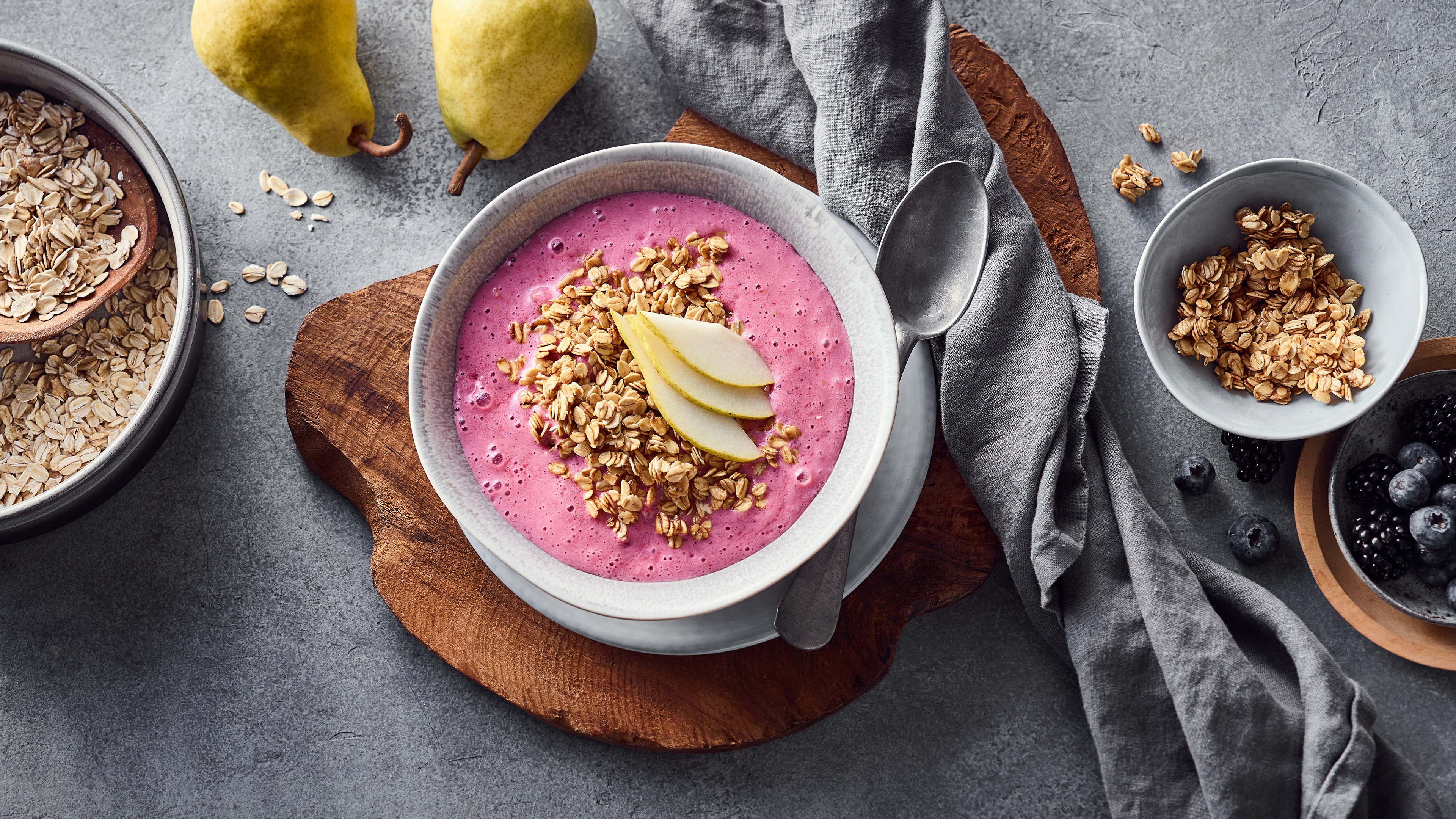 Schüssel mit pinkem Beeren-Smoothie, garniert mit Haferflocken und Birnenscheiben, auf einem Holzbrett; daneben liegen Birnen, Beeren und Müsli.