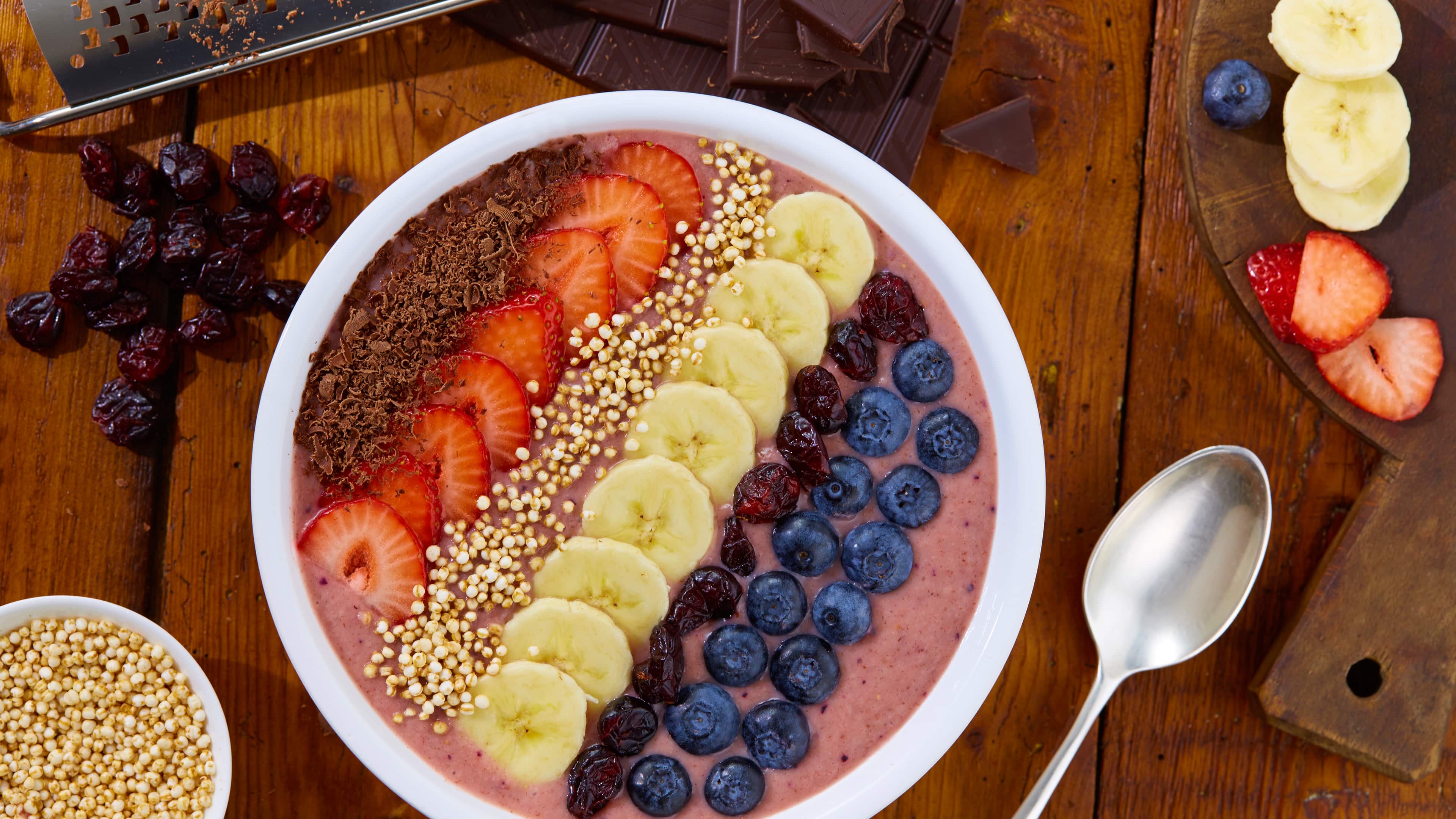 Eine Schale mit cremigem beerenfarbenem Brei, getoppt mit Erdbeeren, Bananen, Blaubeeren, Rosinen, Schokoraspeln