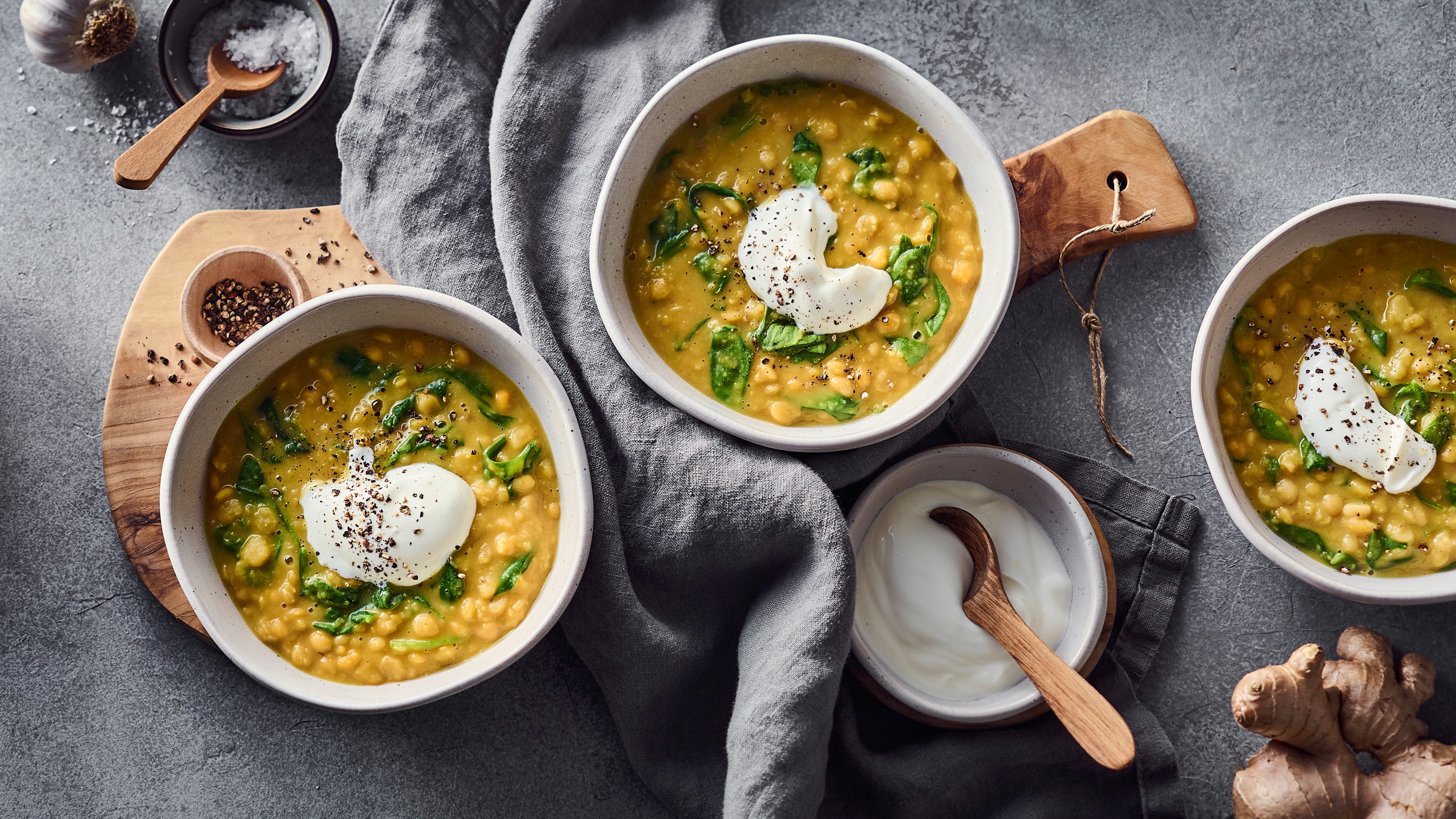Drei Schüsseln mit cremiger gelber Erbsensuppe, garniert mit Joghurt und Blattgemüse, auf einem grauen Tisch mit Gewürzen und frischem Ingwer.