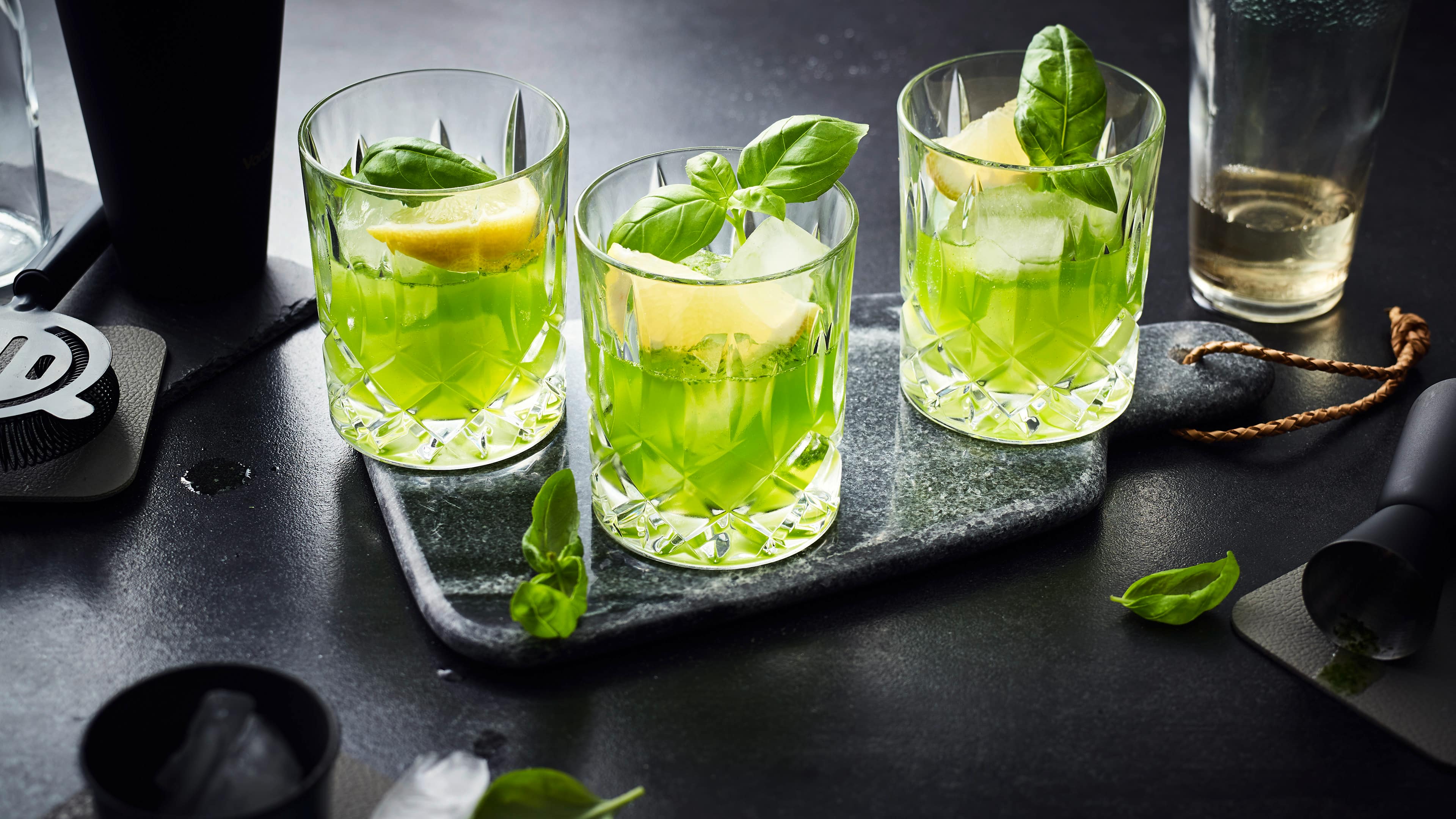 Rezept für Gin Basil Smash Longdrinkgläser mit hellgrünem Getränk, Eiswürfeln, Zitronen-Spalten, getoppt mit frischem Basilikum-Blättchen