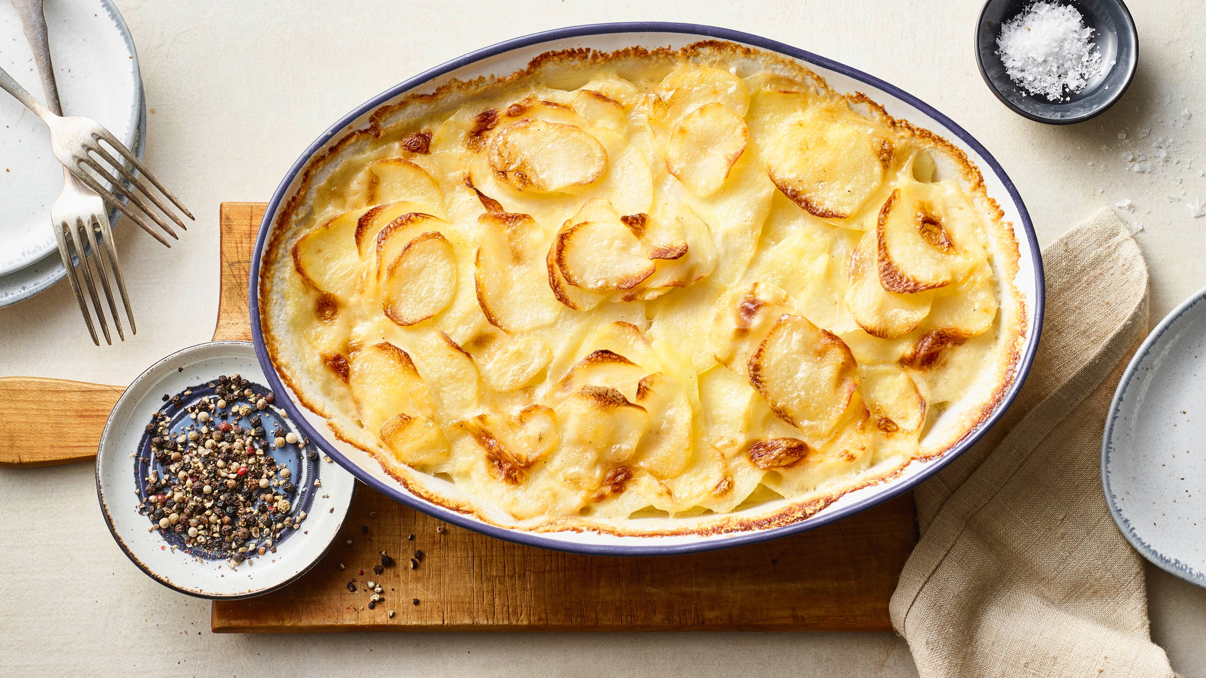 Rezept für französisches Gratin dauphinois Eine Auflaufform mit knusprig überbackenen Kartoffelscheiben, auf einem rustikalen Holzbrett