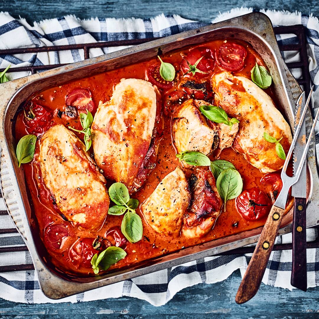 Eine eiserne Auflaufform mit Hähnchen in fruchtiger Tomatensauce und gebackenen Kirschtomaten, getoppt mit Basilikum