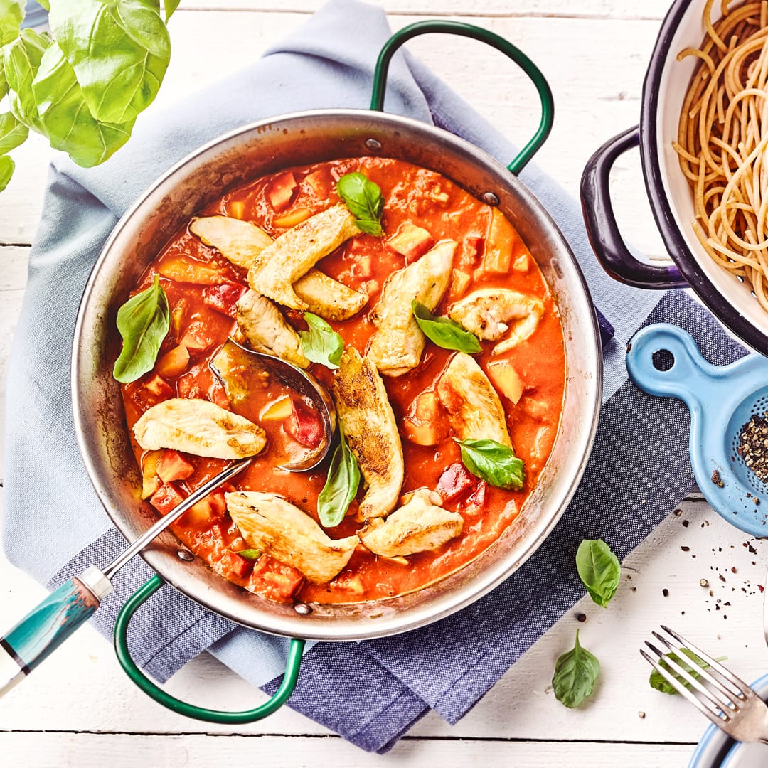 Ein gusseiserner Topf mit Hähnchenstückchen in einer Tomaten-Gemüse-Soße, daneben ein Topf mit Vollkorn-Spaghetti