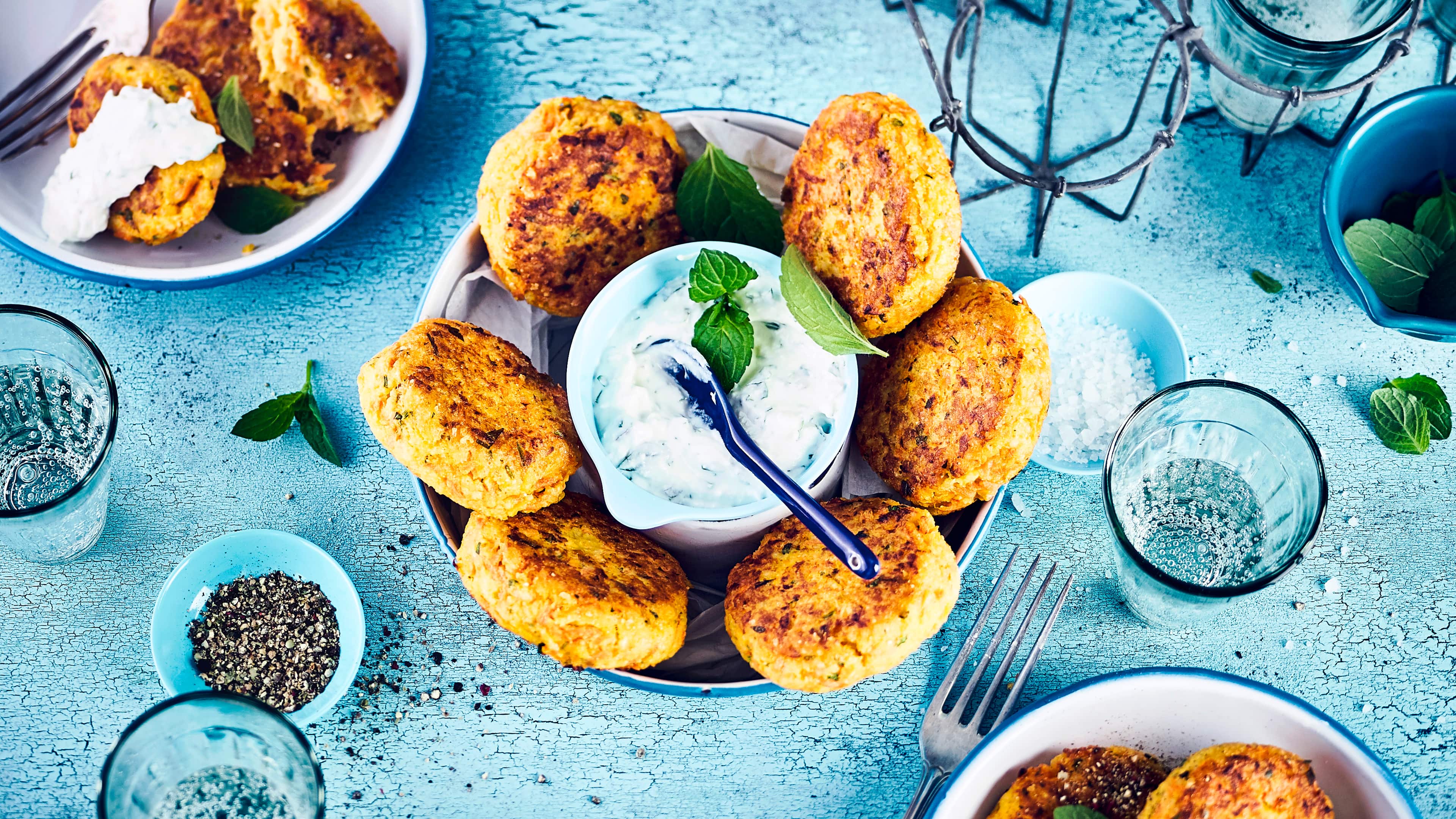 Rezept für herzhafte Hirse-Plätzchen Eine Schüssel mit sechs gelb-braunen, gebratenen Talern, die um eine kleine Schale mit einer weißen Creme angeordnet sind