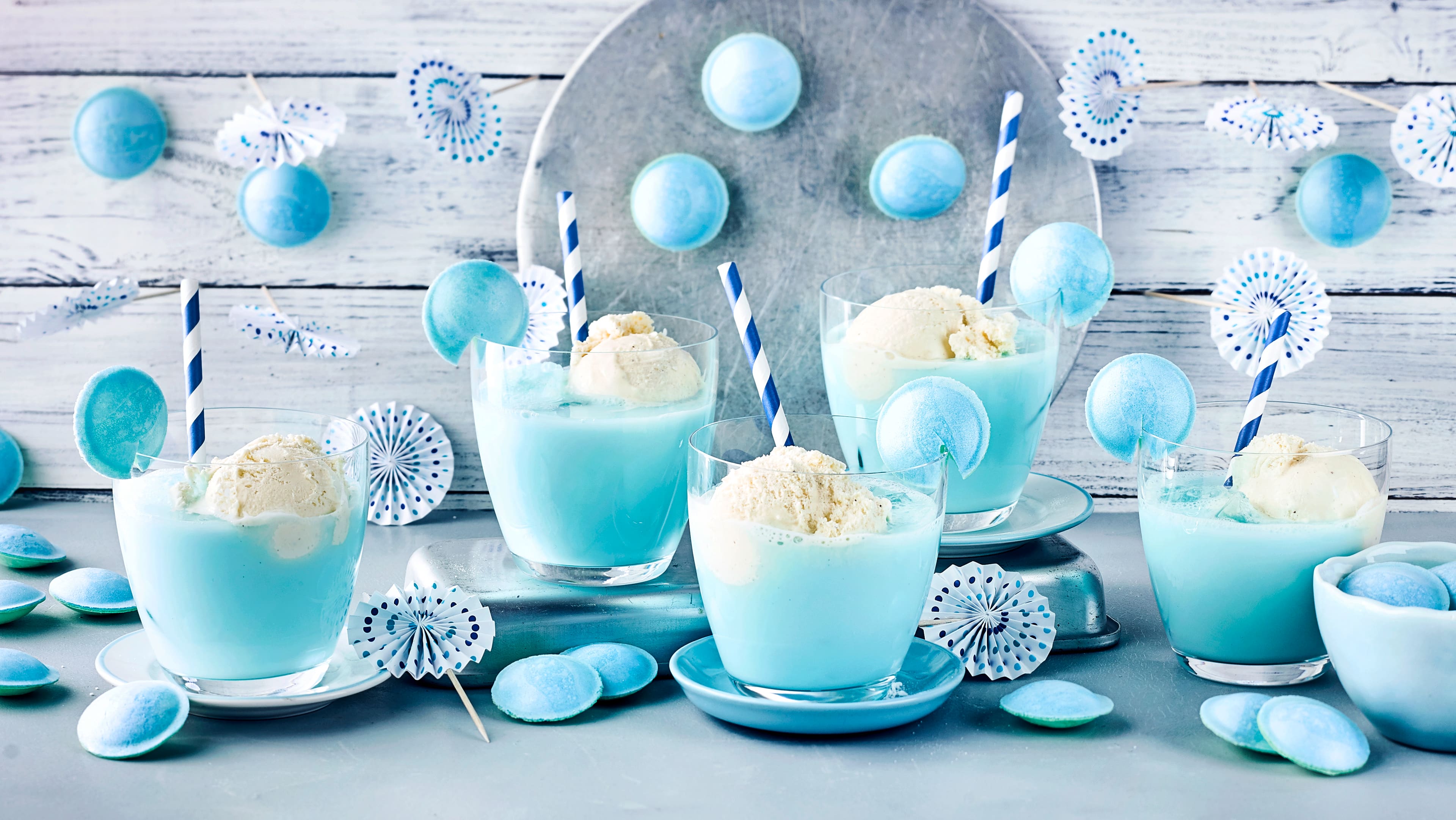 Fünf Gläser mit hellblauer Flüssigkeit, blau-weißen Strohhalmen und einer Kugel Vanilleeis in jedem Glas