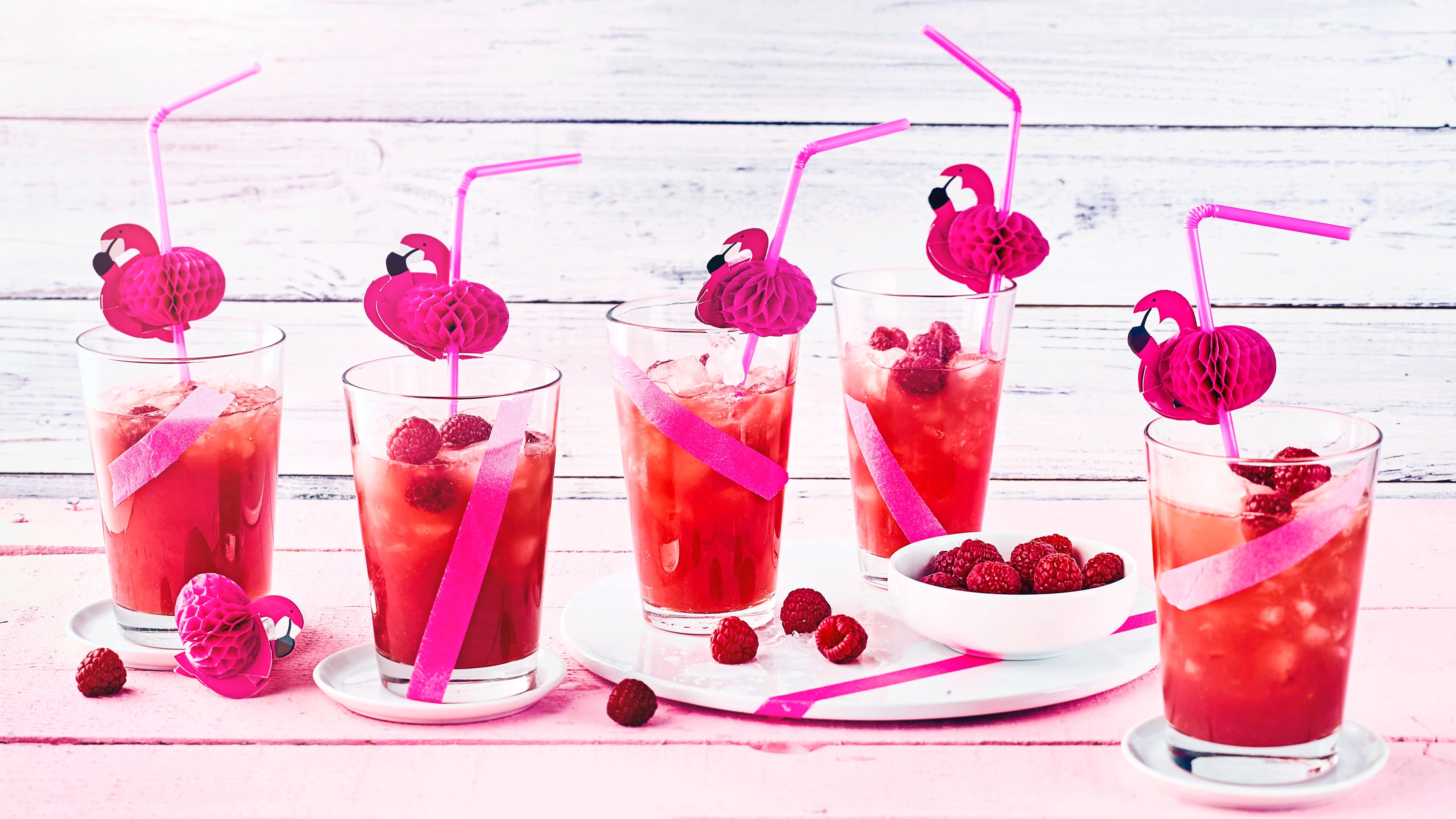 Gläser mit einer rötlichen Flüssigkeit, Eiswürfeln und Himbeeren, dekoriert mit pinken Strohhalmen und Papier-Flamingos