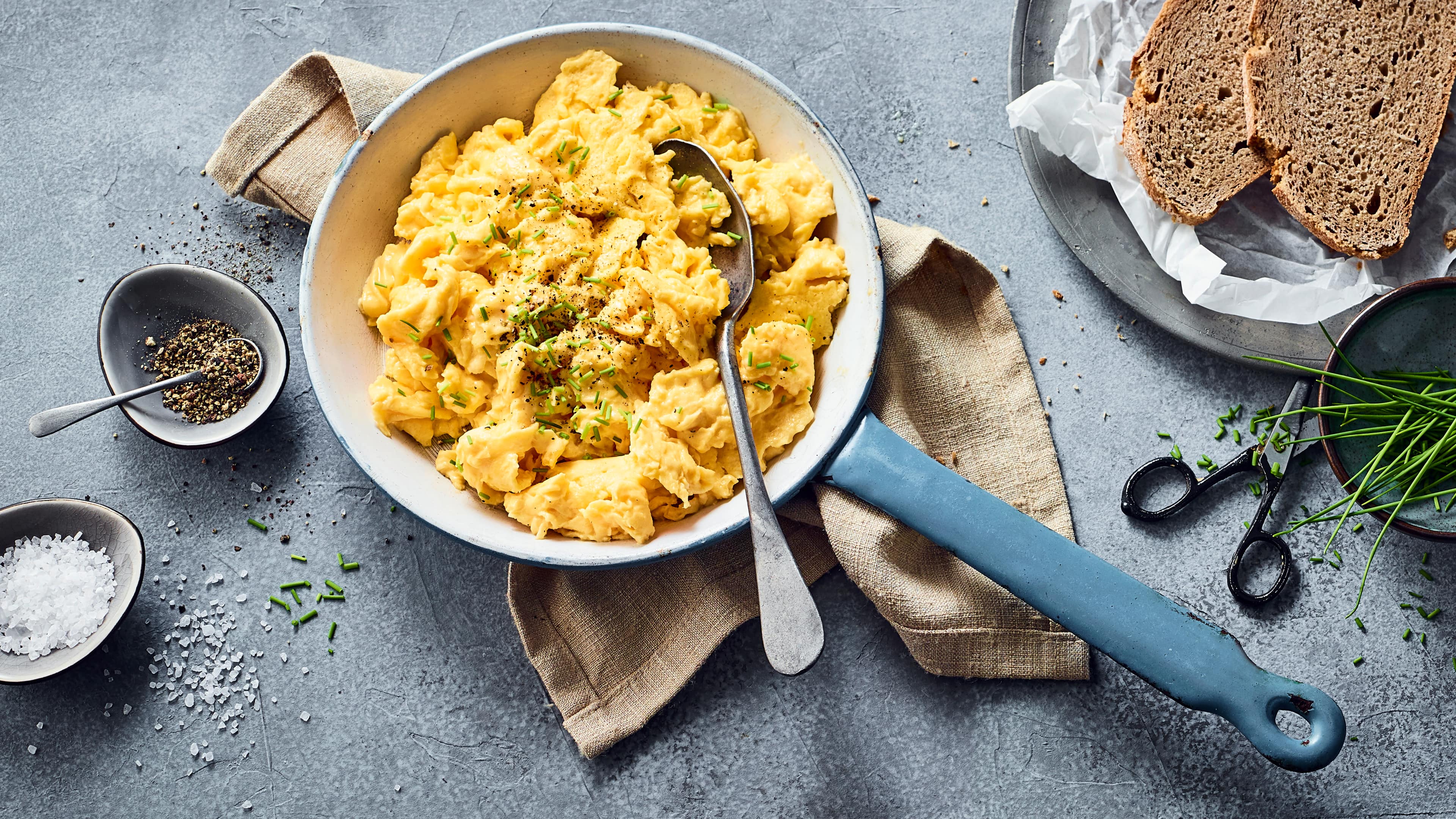 Rezept für klassisches Rührei Eine gusseiserne Pfanne mit fluffigem Rührei, getoppt mit frischem Schnittlauch, daneben ein Schälchen mit Pfeffer