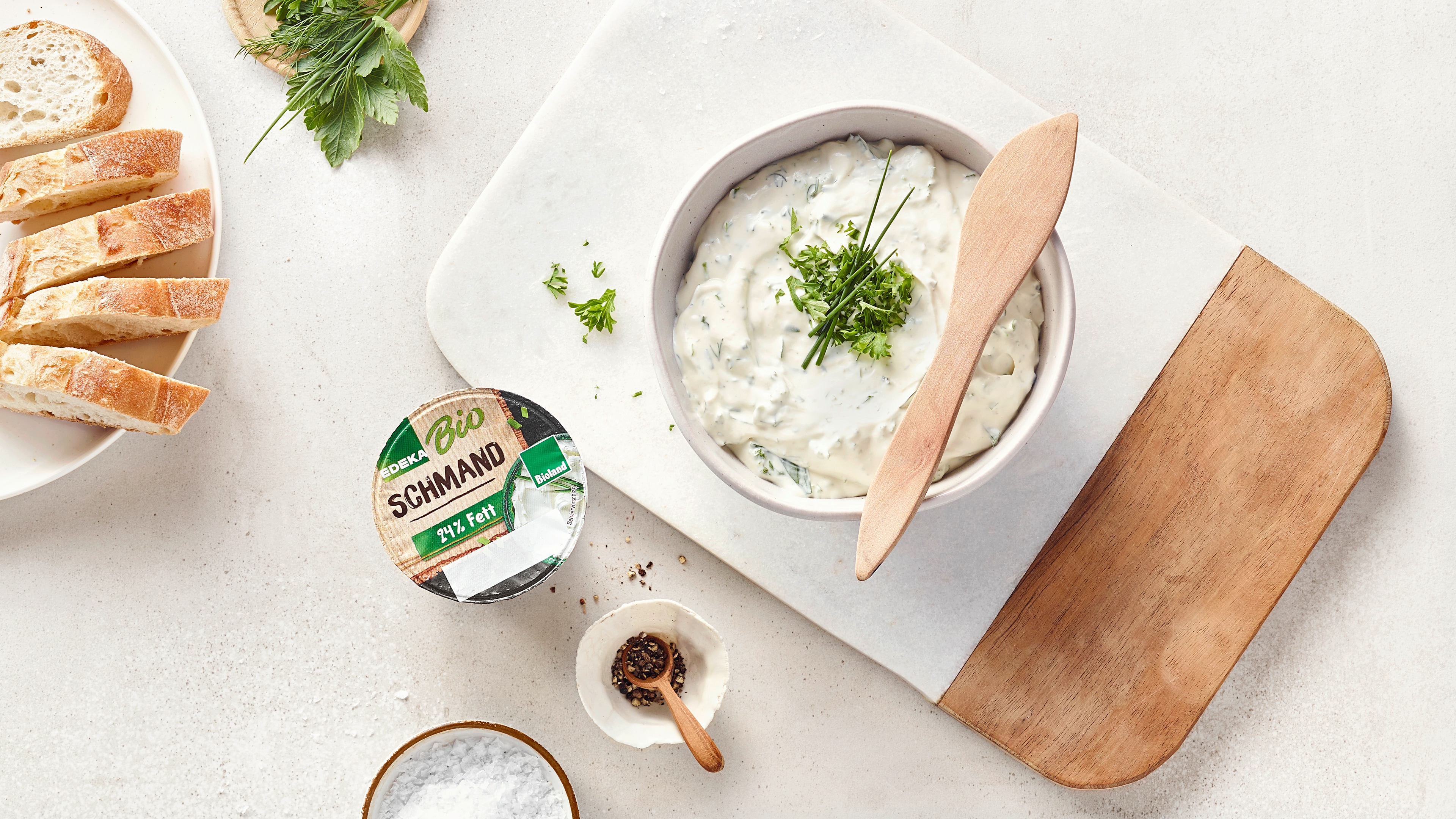 Schüssel mit cremigem Kräuterdip auf einem Holzbrett, daneben Brot, Gewürze und ein Becher EDEKA Bio Schmand.
