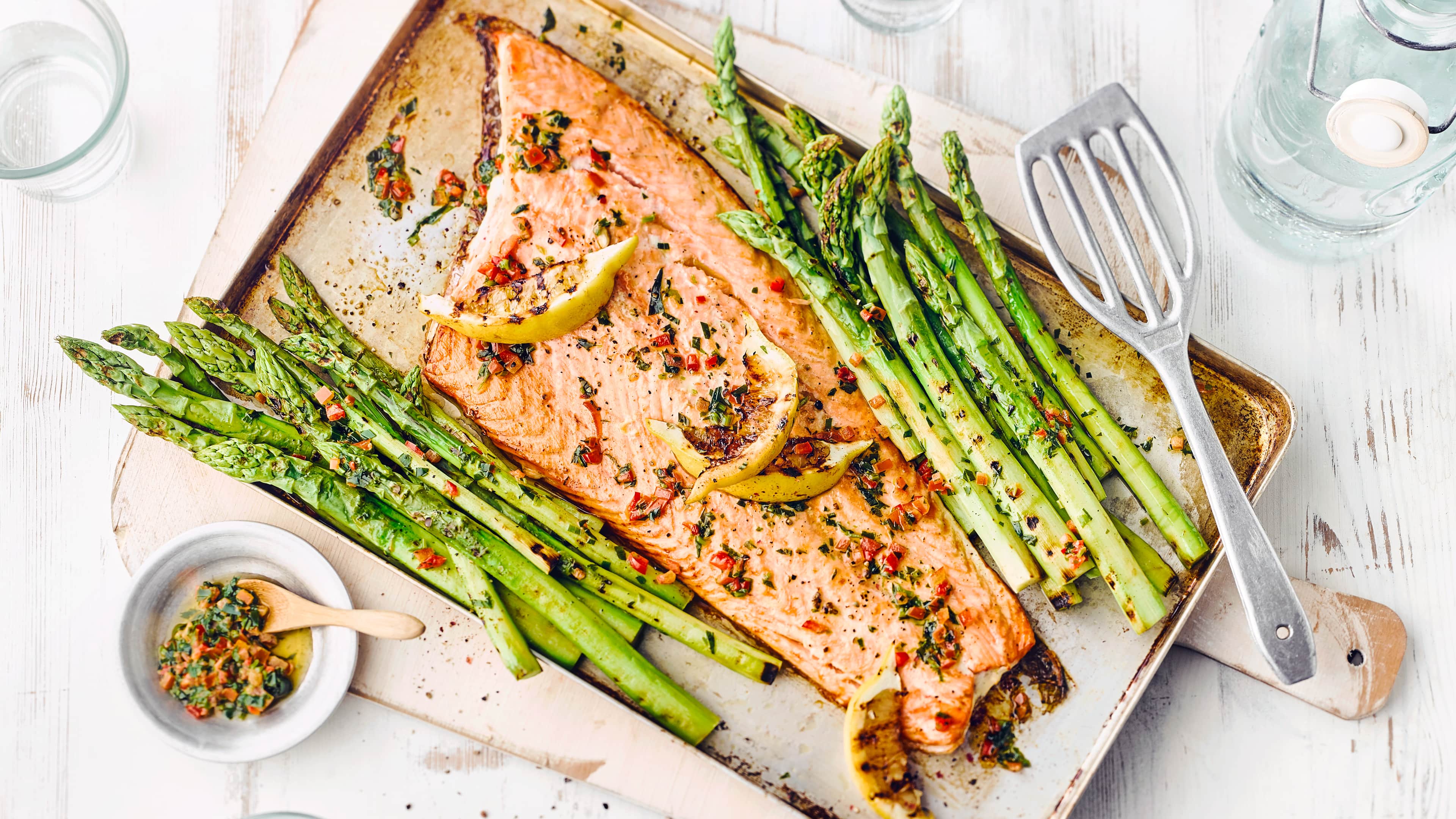 Rezept für Lachs mit grünem Spargel Ein Blech mit gebratenem Lachs und grünem Spargel, getoppt mit Zitronenspalten, daneben ein Schälchen mit Kräuterbutter