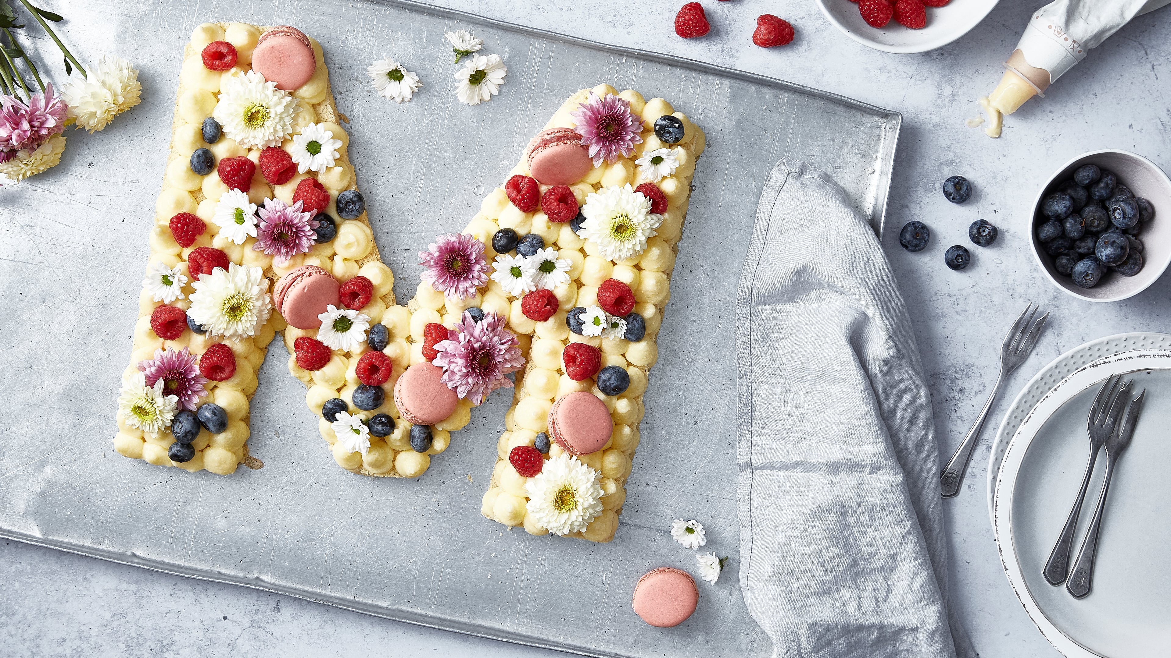 Rezept für Letter Cake Ein Kuchen in Form des Buchstaben M, dekoriert mit Creme-Tupfen, frischen Himbeeren, Blaubeeren, Blumen und pinken Macarons