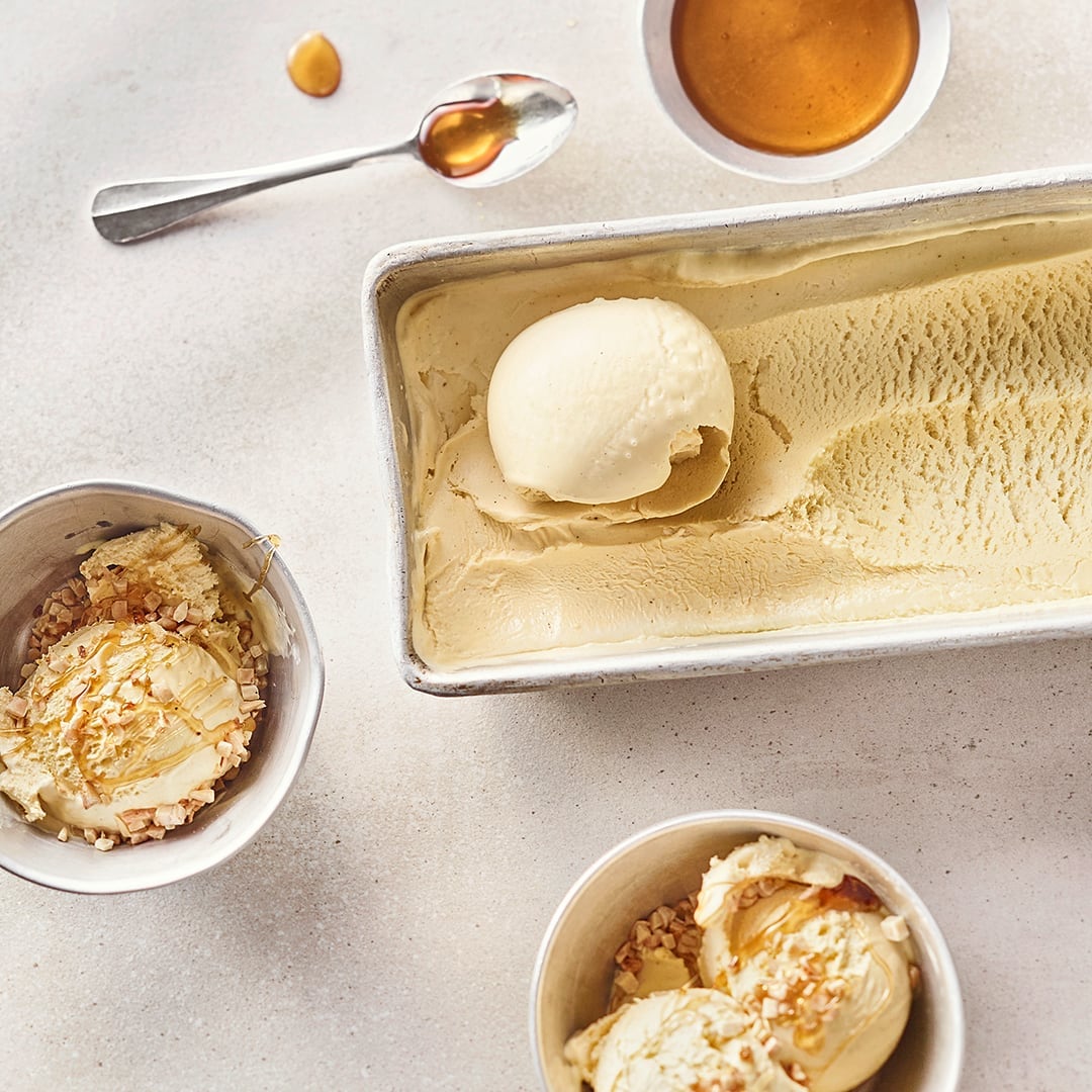 Ein metallischer Eisbehälter und zwei kleine Schüsseln, gefüllt mit cremefarbenen Eis und getoppt mit Honig und Mandeln