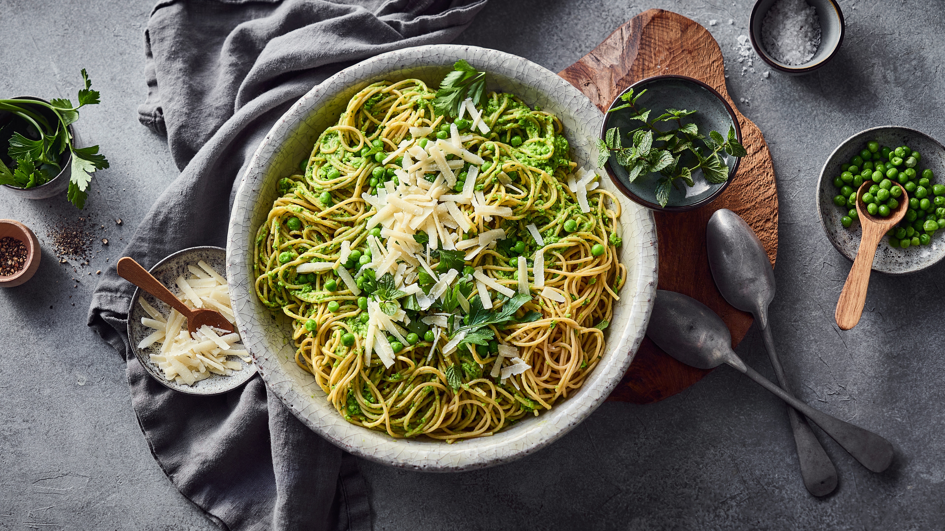 Schüssel mit Spaghetti in grünem Kräuterpesto, garniert mit Erbsen und gehobeltem Käse, umgeben von frischen Kräutern auf einem grauen Tisch.