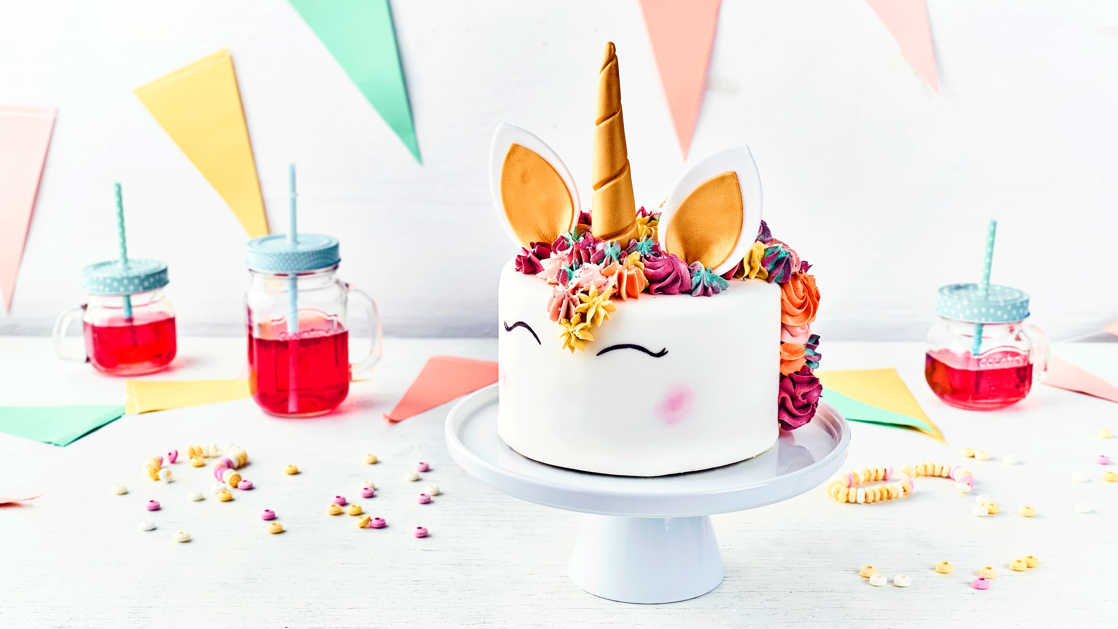 Rezept für eine Einhorn-Motivtorte Eine weiße Einhorn-Tore mit bunkter Buttercreme-Mähne, goldenem Horn und Fondant-Ohren, auf einer weißen Etagere