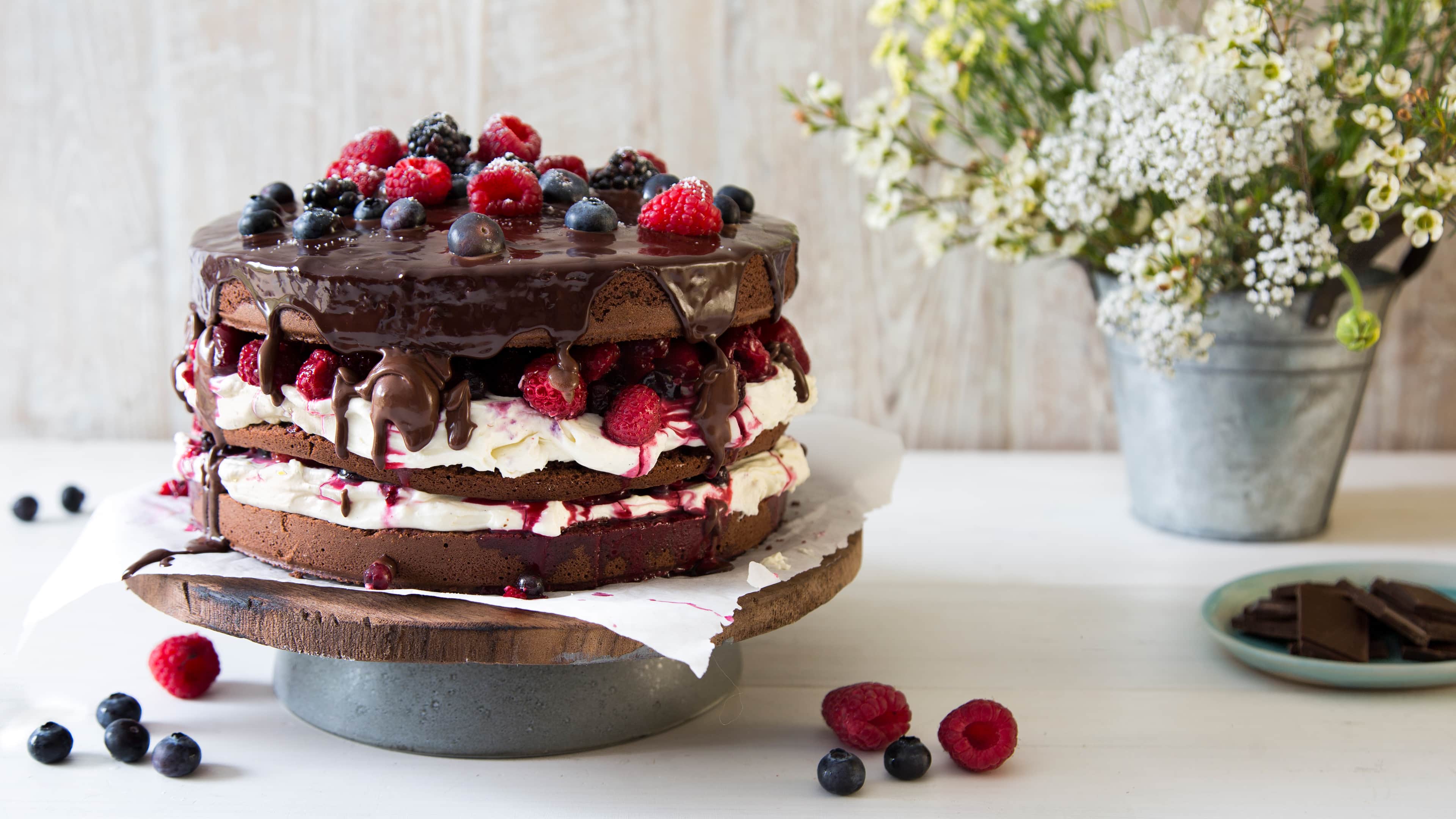 Rezept für Naked Chocolate Cake mit Beeren Eine Torte mit Schokoböden, weißer Creme, frischen Früchten und Schokoladen-Drip, auf einer Holzetagere