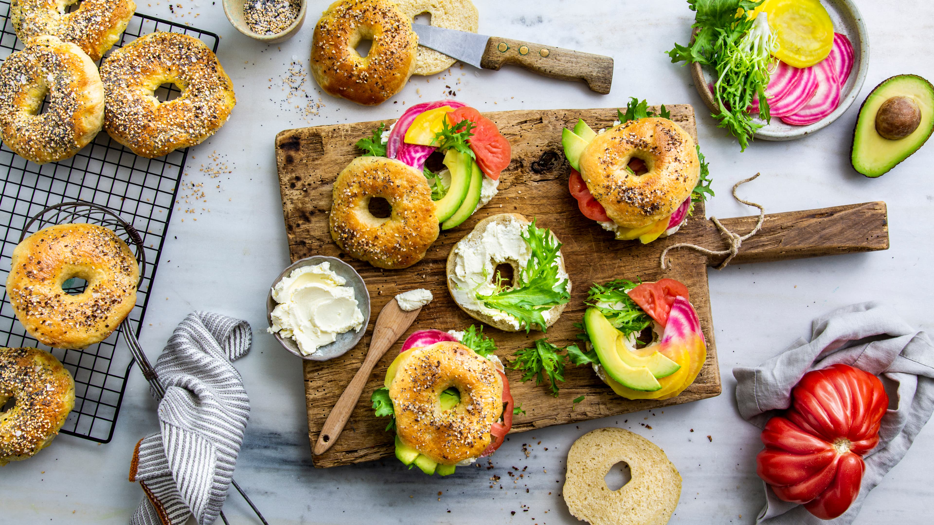 Rezept für New York Style Bagel Bagels mit Mohn und Sesam, belegt mit cremigem Frischkäse, Avocado, Salat, Tomate und Ringelbete, auf einem Holzbrett
