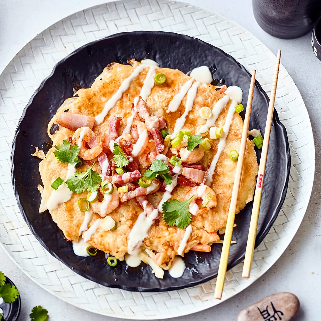 Okonomiyaki kommen aus Japan und sind eine Art dünner Pfannkuchen, der mit einer typischen Okonomiyaki-Sauce gewürzt und mit Speck, Zwiebeln und Garnelen belegt wird.