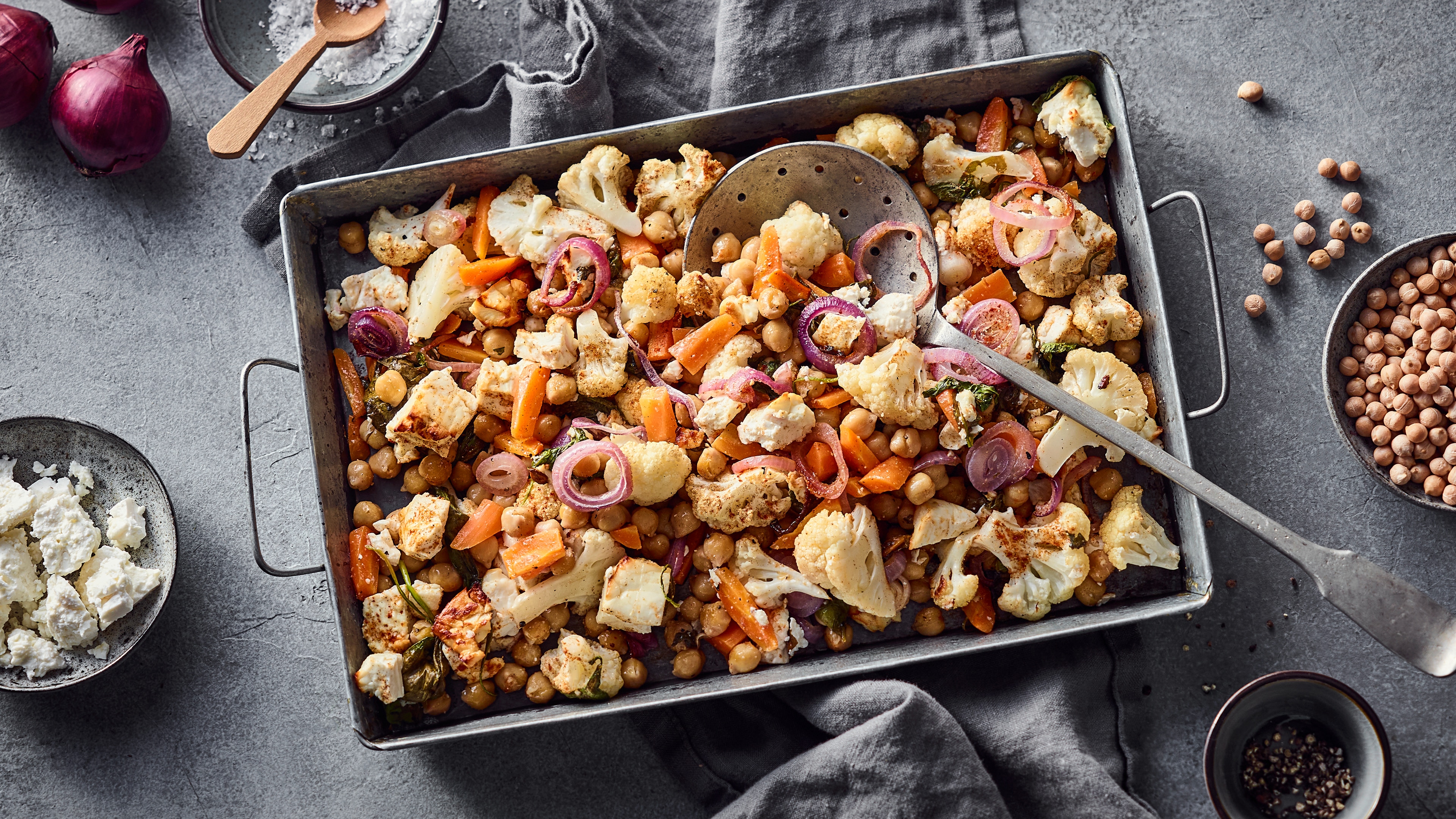 Ofengemüse mit Blumenkohl, Kichererbsen, Karotten, roten Zwiebeln und Feta in einer Backform, serviert auf einem grauen Tisch mit Gewürzen und frischen Zutaten.