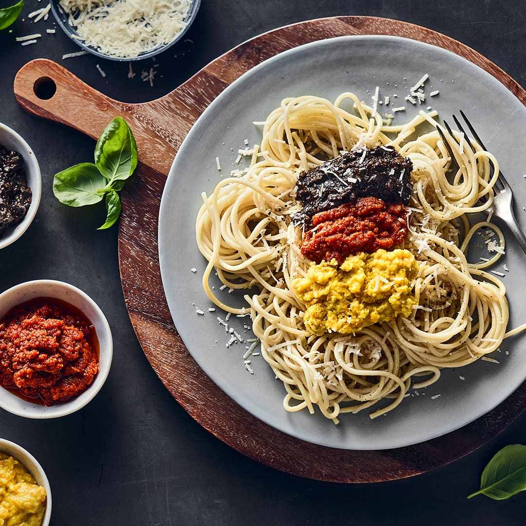 Ein runder Teller mit Spaghetti, getoppt mit schwarzen, roten und gelben Pesto sowie Parmesan