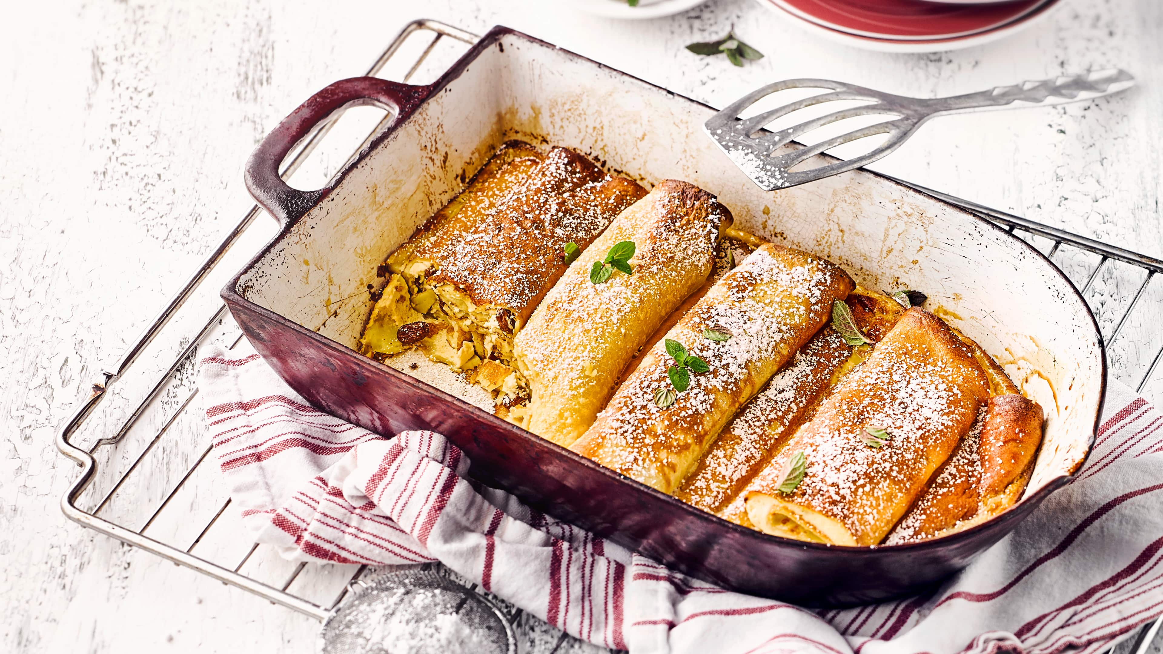 Rezept für Pfannkuchen-Auflauf Eine rustikale Auflaufform mit gelbgoldenen Teigrollen mit Rosinen, bestreut mit Puderzucker