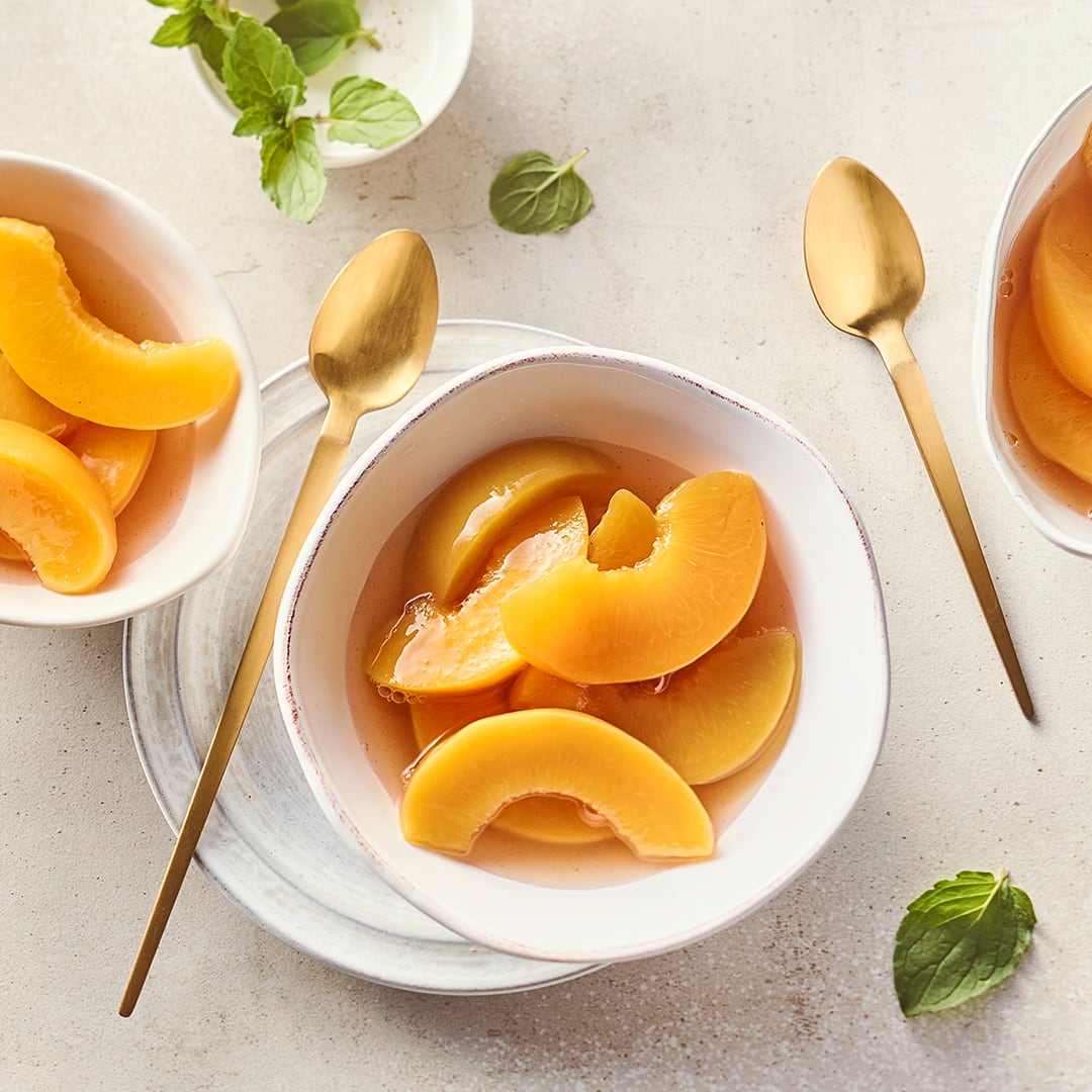Kleine Schüsseln befüllt mit einem Kompott aus Pfirsichscheiben und Fruchtsaft, daneben goldene lange Dessertlöffel