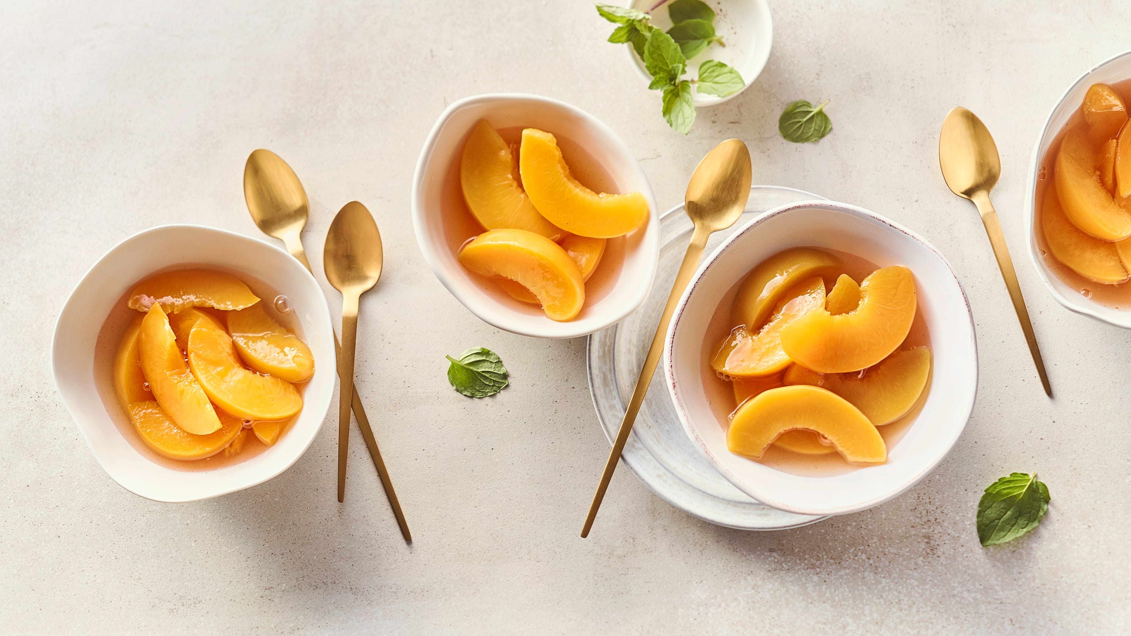 Rezept für Pfirsichkompott Kleine Schüsseln befüllt mit einem Kompott aus Pfirsichscheiben und Fruchtsaft, daneben goldene lange Dessertlöffel