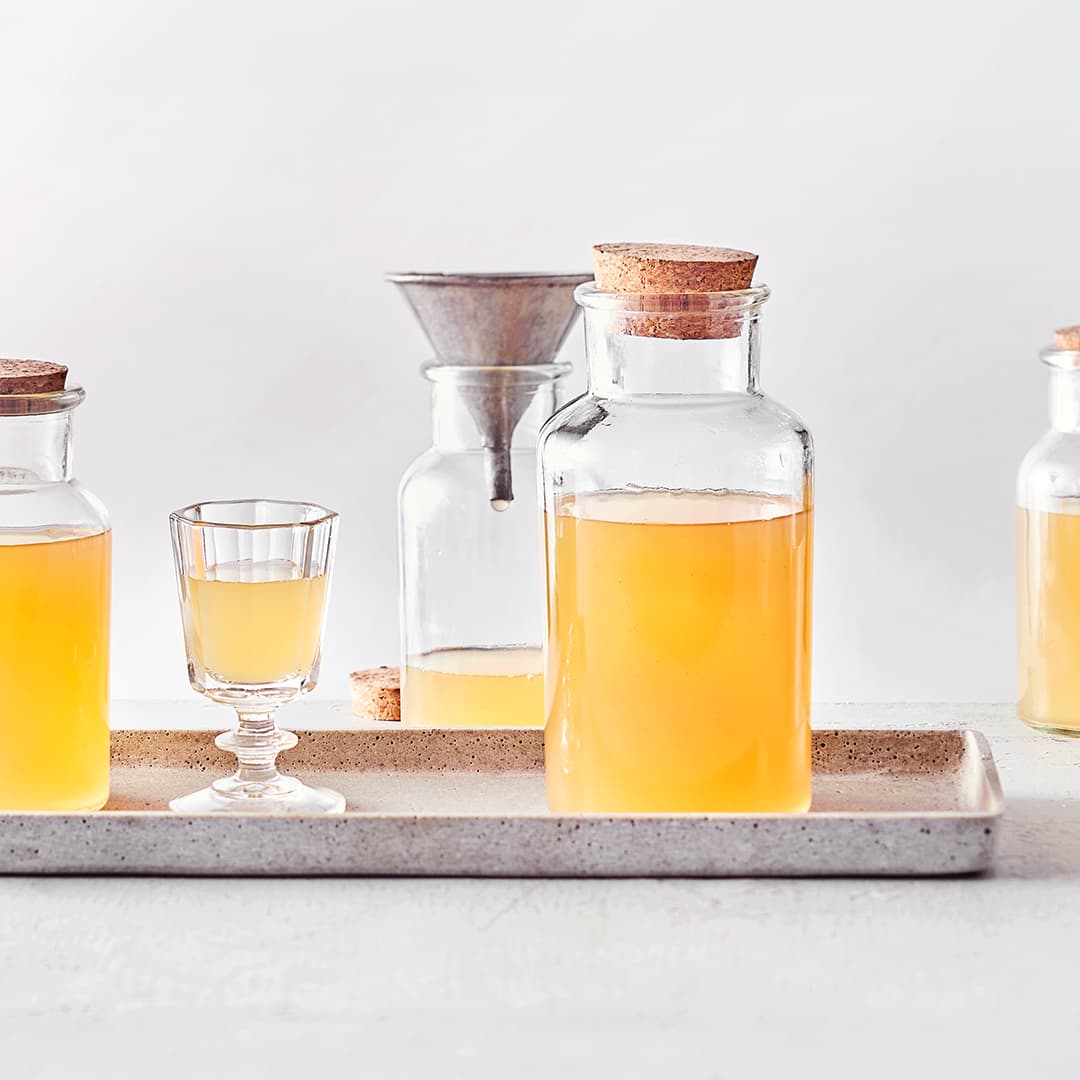 Zwei verschlossene Flaschen mit Korken und ein eckiges Trinkglas, befüllt mit orange-gelbem klarem Likör