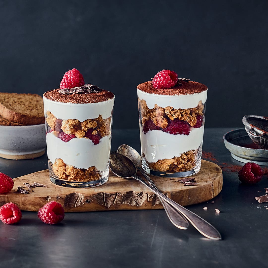 Dessertgläser mit Tiramisu aus Schichten von Creme, Himbeeren und Crumble, bestreut mit Kakao und Zartbitterschokolade