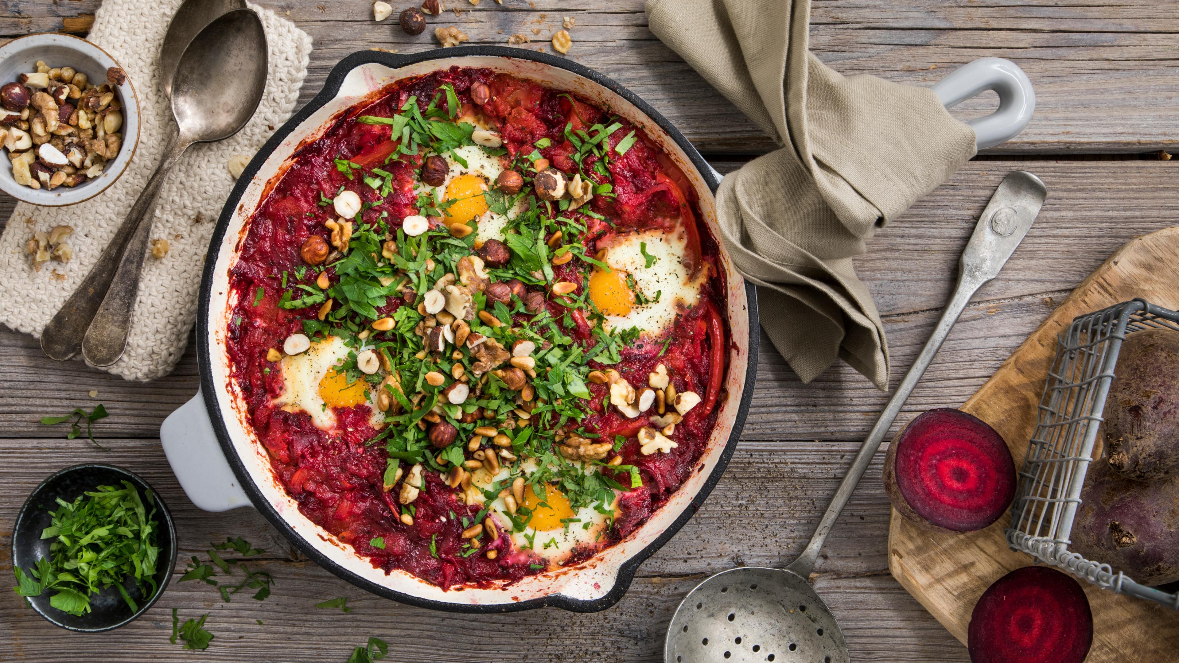 Rezept für Rote Bete Shakshuka Eine große Pfanne mit cremigem, rot-pinken Inhalt, darin vier pochierte Eier und Toppings aus Nüssen, Kernen und Kräutern