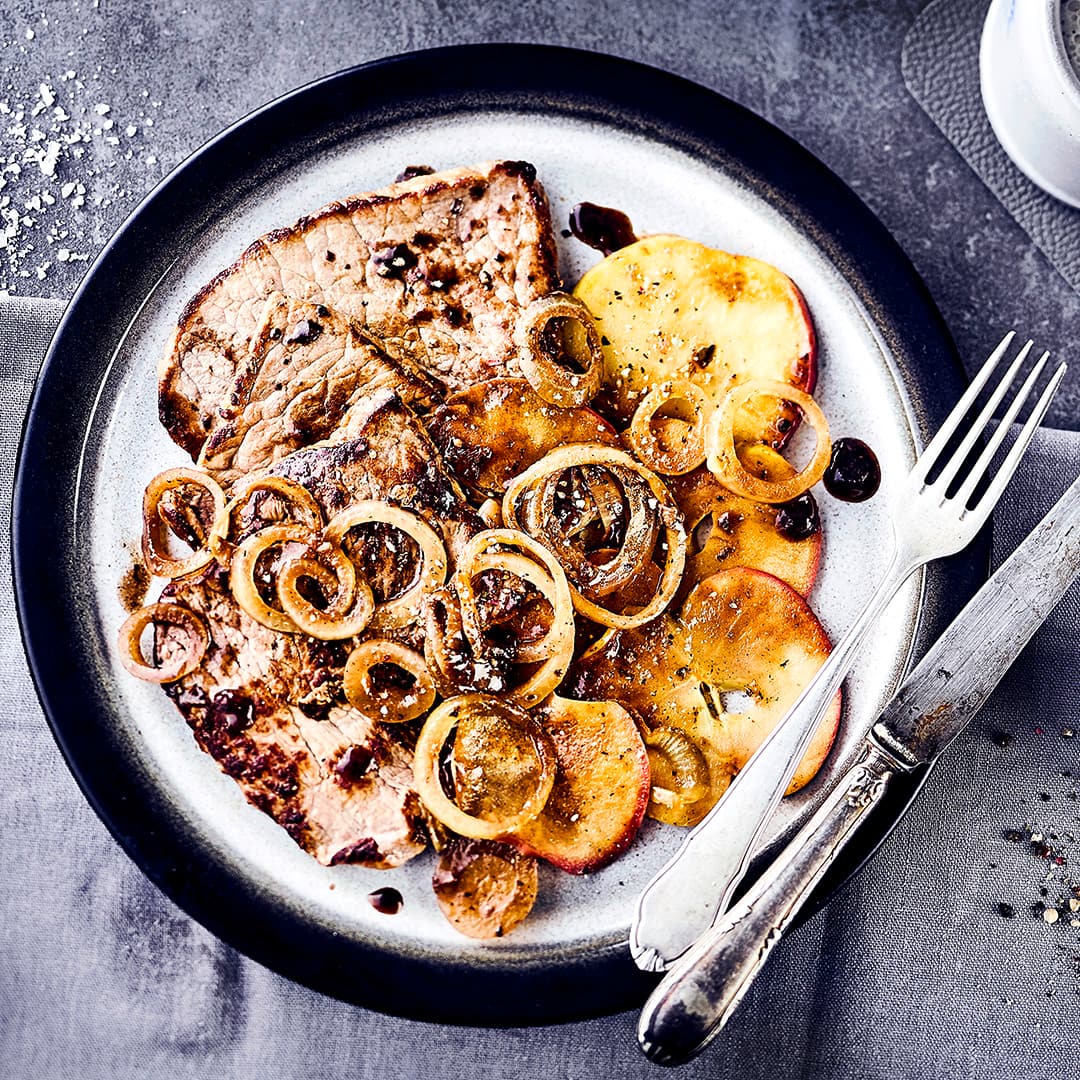 Ein Teller mit knusprig gebratenem Fleisch, Kartoffelscheiben, getoppt mit karamellisierten Zwiebelringen und dunkler Soße