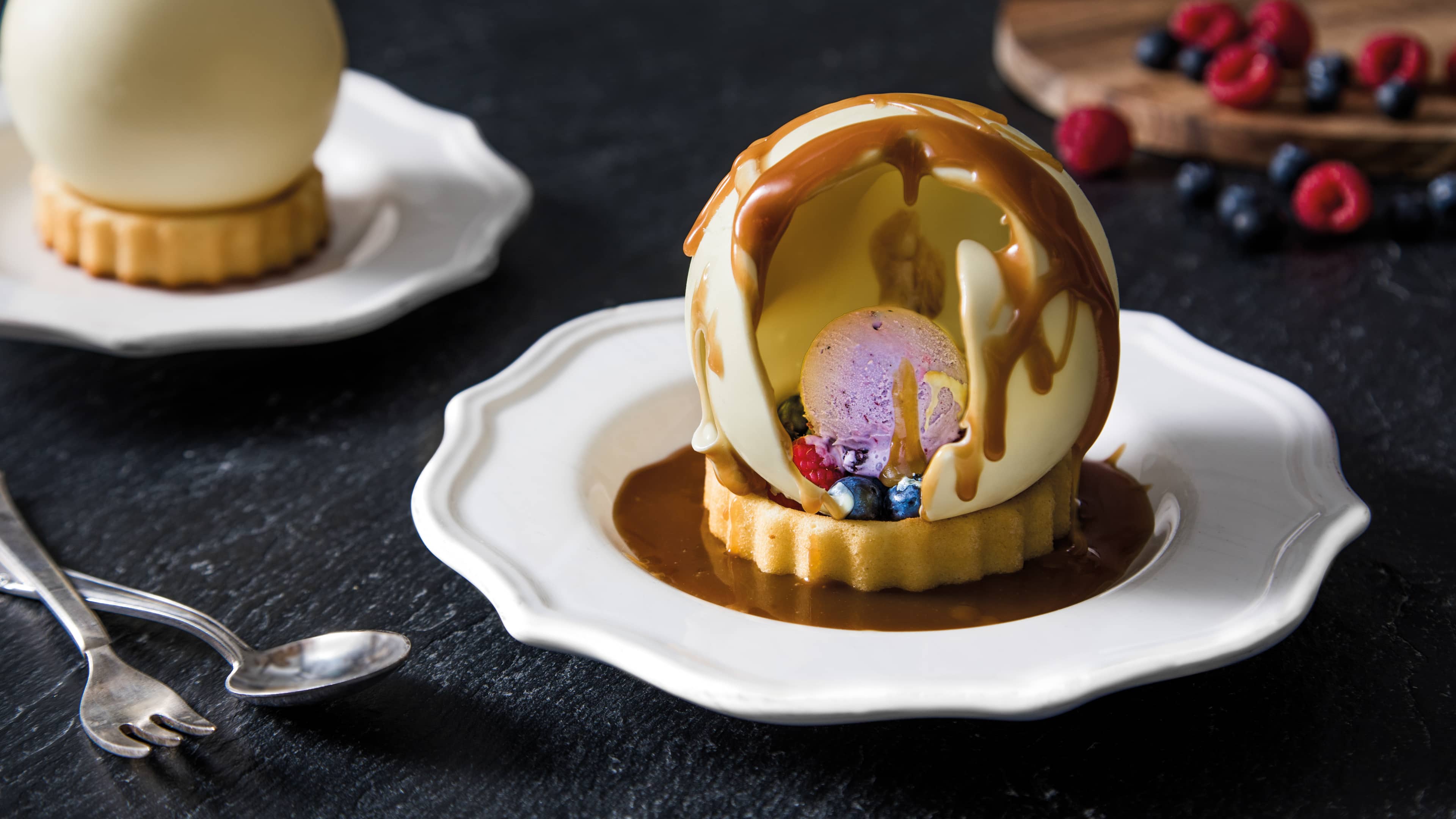 Rezept für Schokokugel-Dessert mit Heidelbeer-Eiscreme und Karamellsoße Ein Tartelette, getoppt mit einer Kugel lila Eiscreme, Beeren, einer weißer Schokoladenhaube und Karamellsoße, auf einem Teller