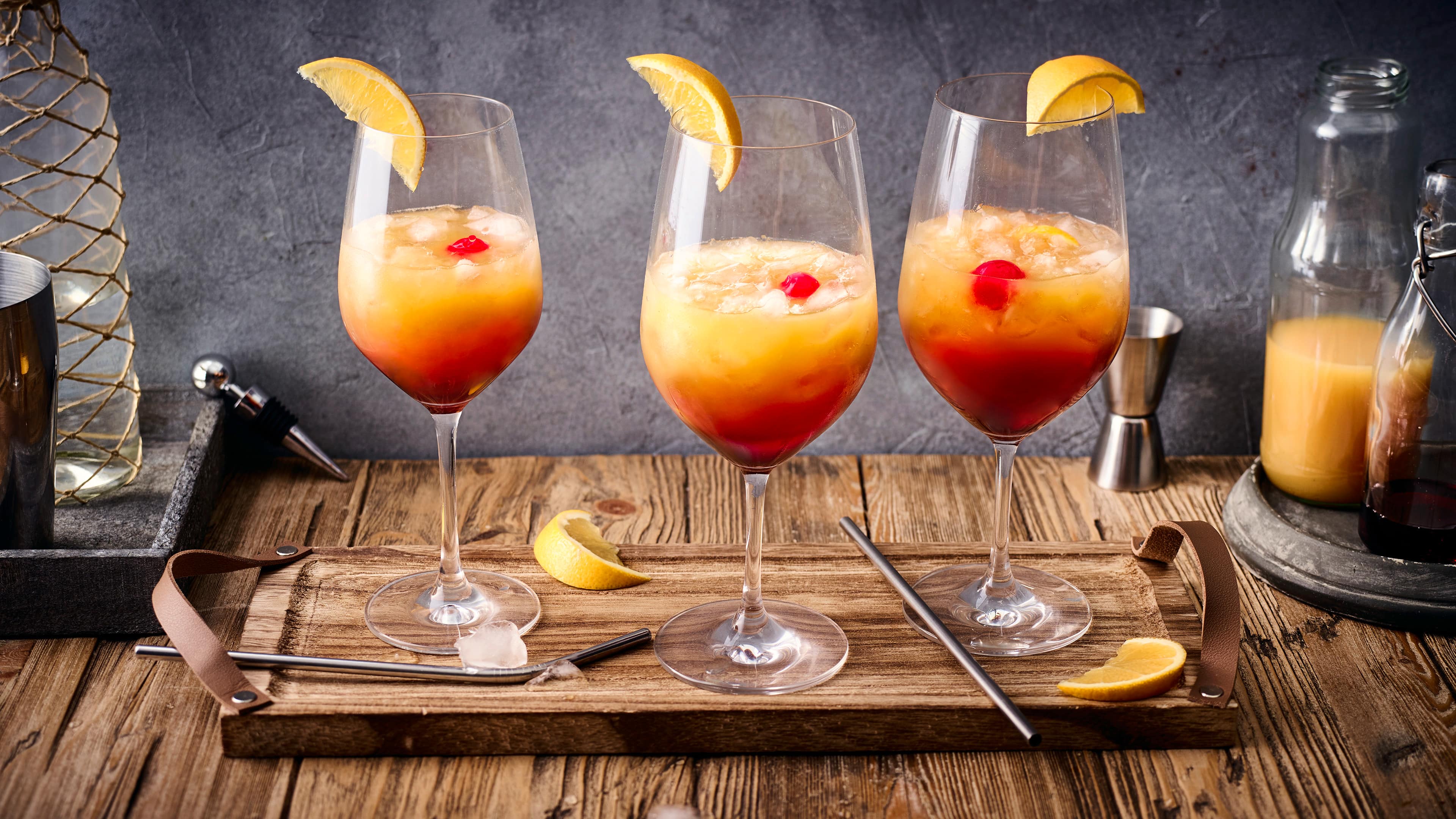 Cocktail-Rezept für Sex on the Beach Drei bauchige Gläser gefüllt mit roter und orangener Flüssigkeit sowie Eiswürfeln, auf einem Holzbrett