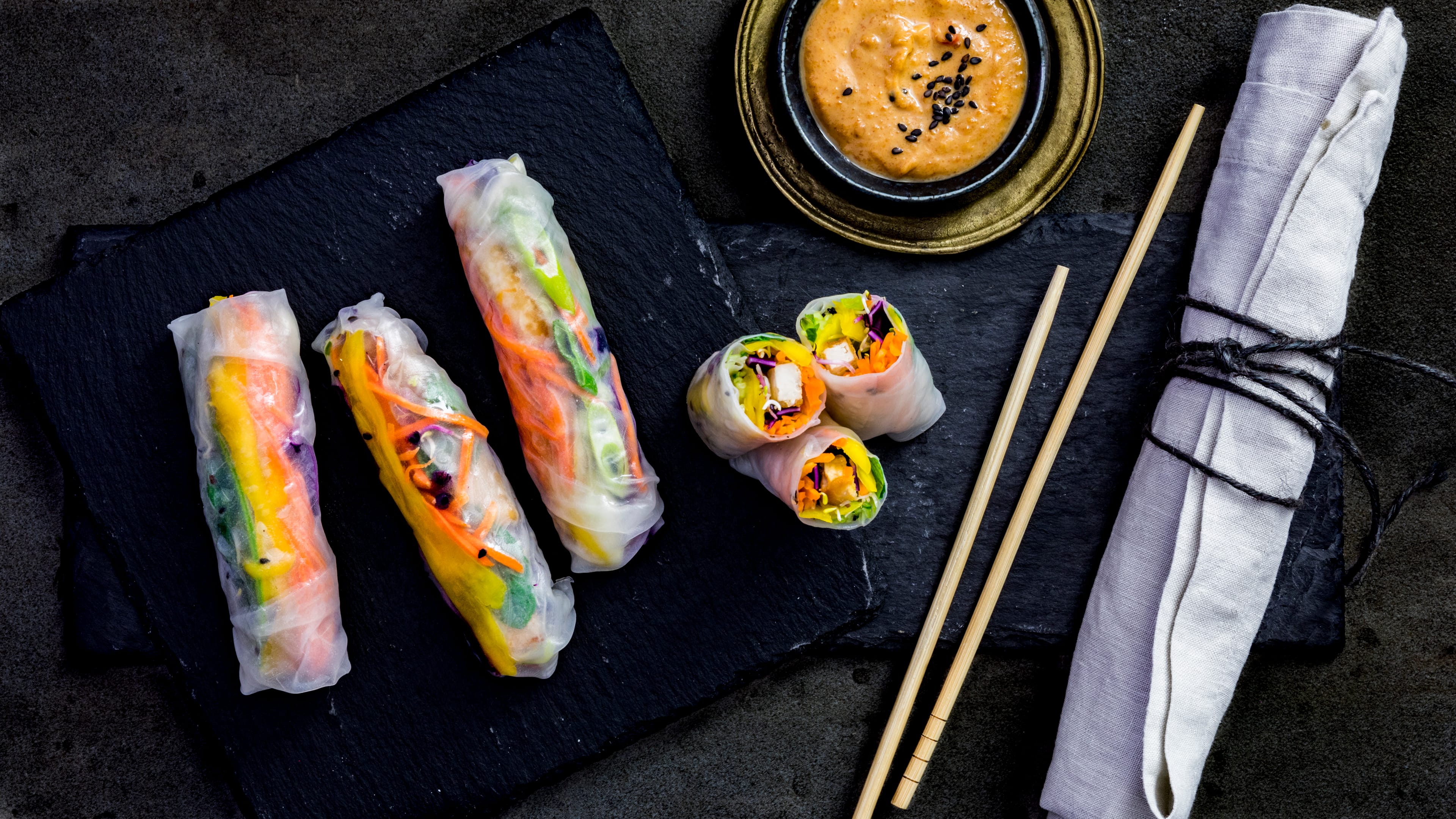 Rezept für asiatische Sommerrollen Reispapier-Rollen, gefüllt mit bunten Gemüse-Sticks und Tofu, auf einem Schiefer-Brett, daneben eine Schale mit cremigem Dip