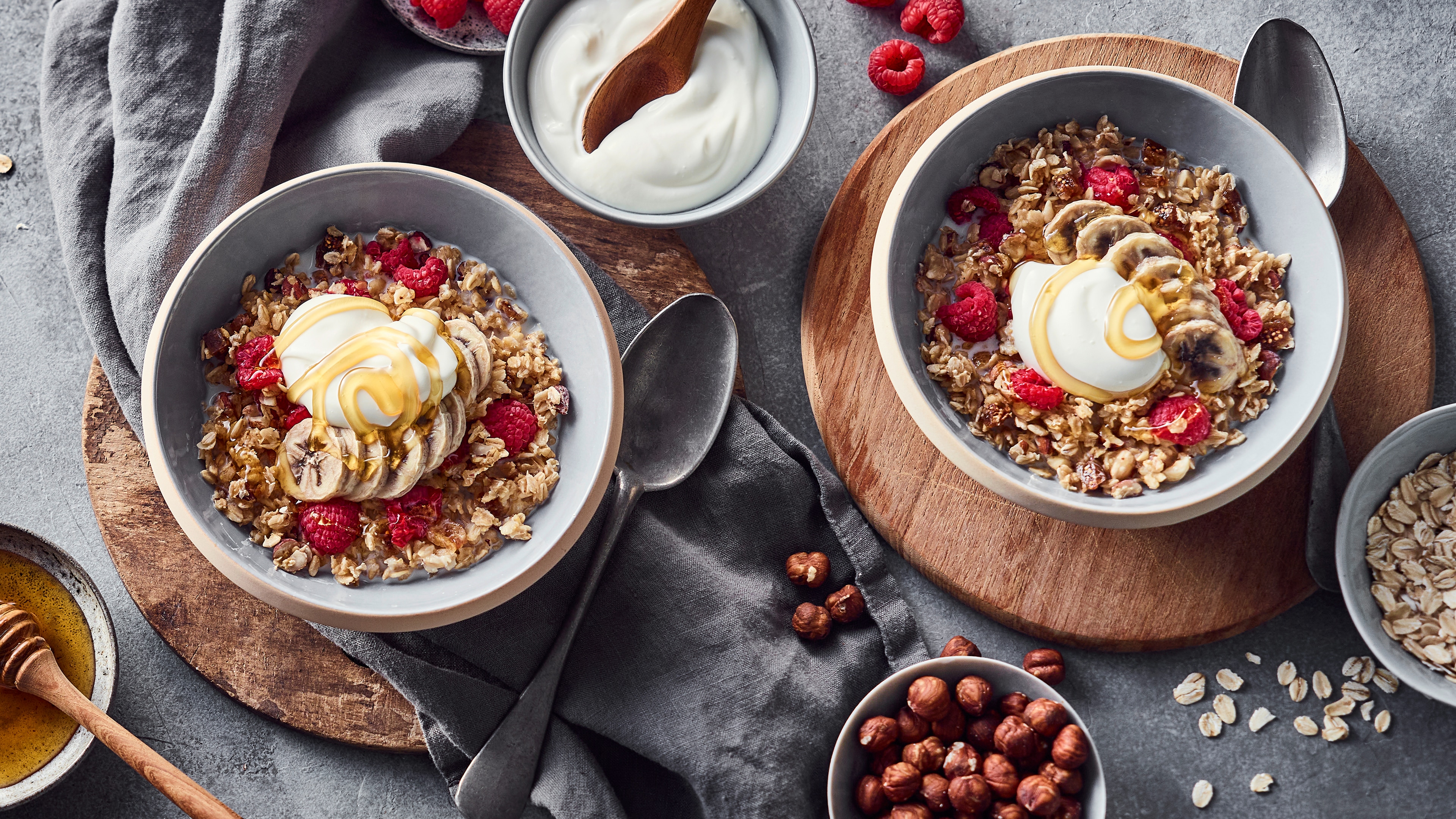 Zwei Schüsseln Haferbrei mit Bananenscheiben, Himbeeren und Joghurt, garniert mit Honig; umgeben von Haferflocken und Haselnüssen.