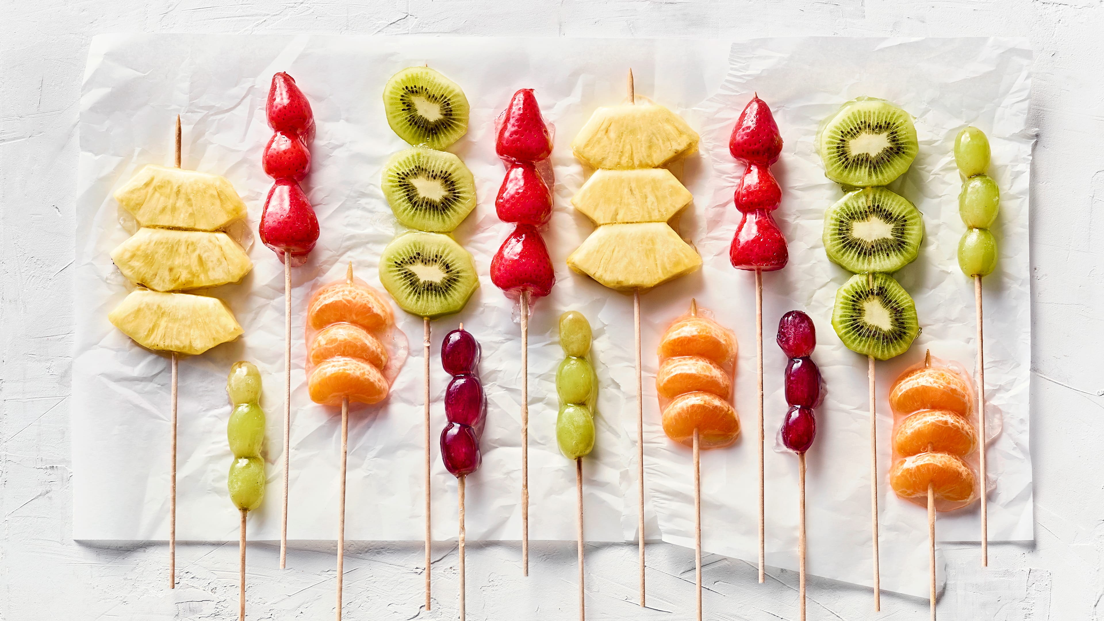 Asiatisches Streetfood-Rezept für Tanghulu Kandierte Kiwi-Scheiben, Erdbeeren, Ananasstückchen und Mandarinenspalten an Holzspießen auf Backpapier