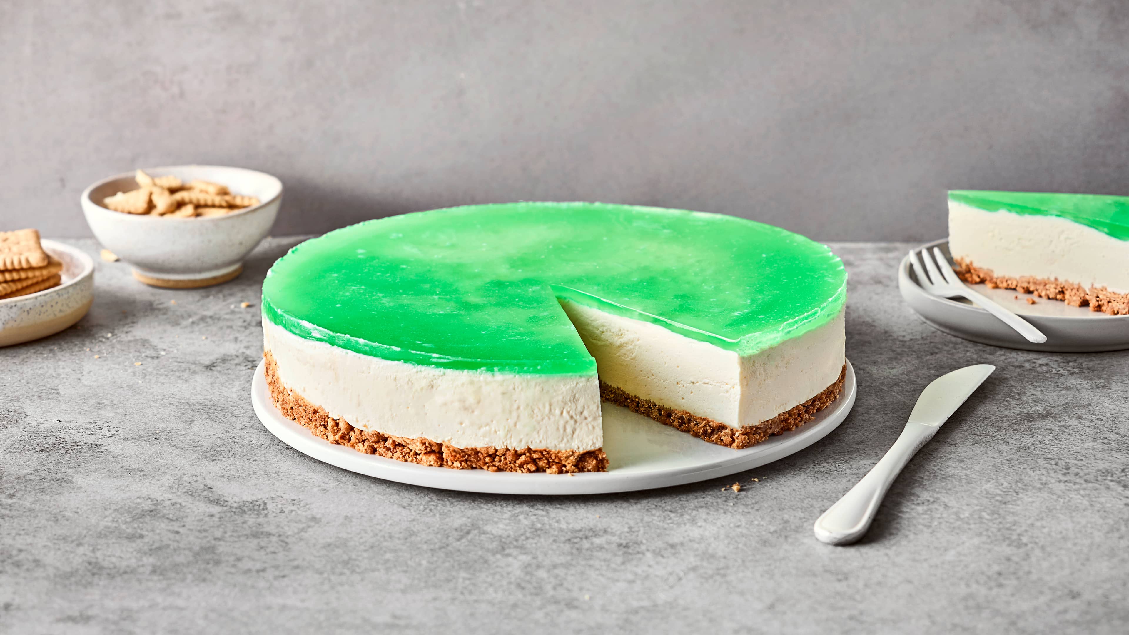 Rezept für Wackelpudding-Kuchen Runde Frischkäsetorte mit einem Boden aus Butterkeks und einer grünen Waldmeister-Wackelpuddingschicht auf einem Teller
