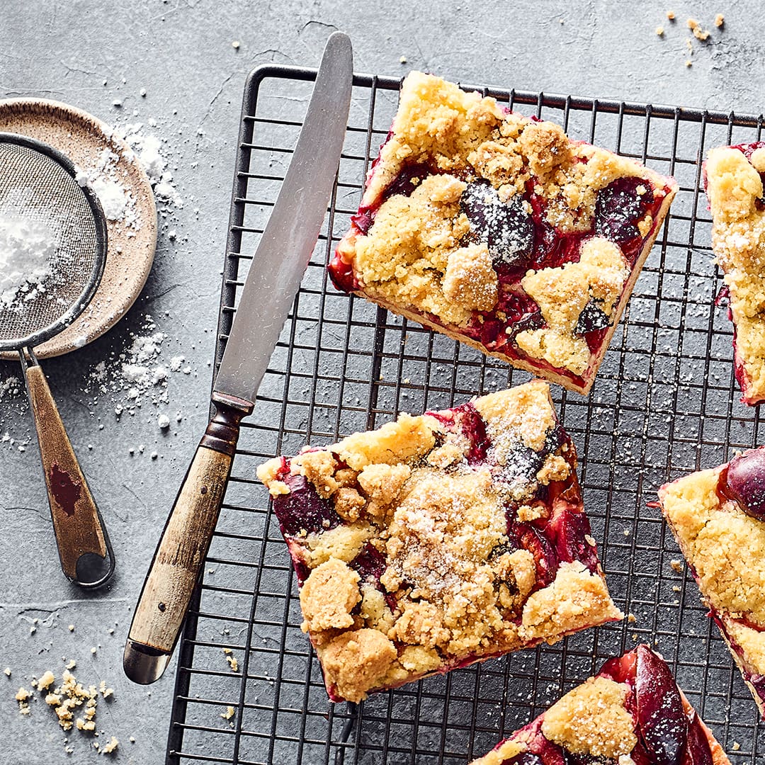 Stücken Blechkuchen mit Zwetschgen und Butterstreuseln, bestreut mit Puderzucker, auf einem Abkühlgitter