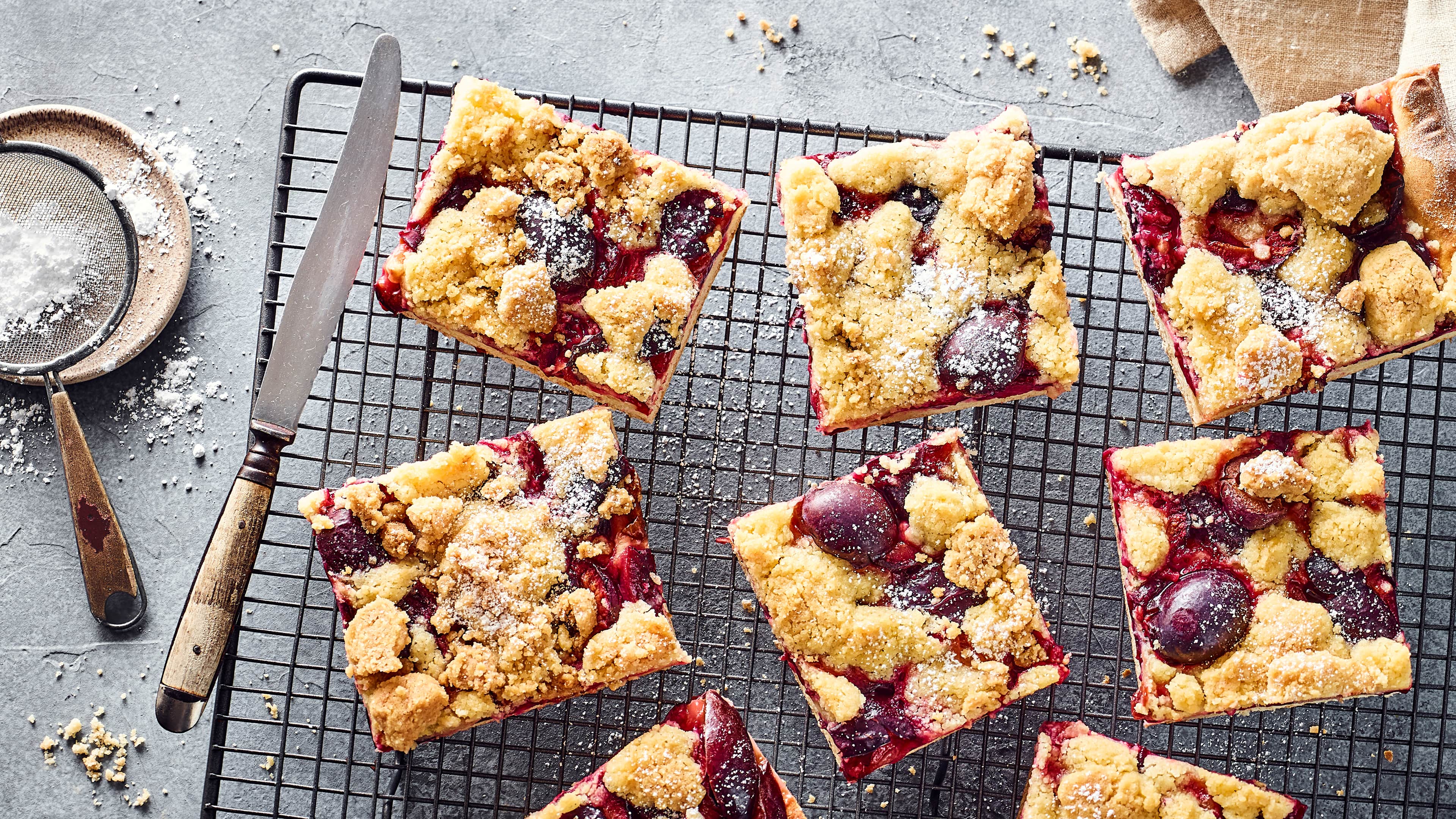 Rezept für Zwetschgen-Streuselkuchen Stücken Blechkuchen mit Zwetschgen und Butterstreuseln, bestreut mit Puderzucker, auf einem Abkühlgitter