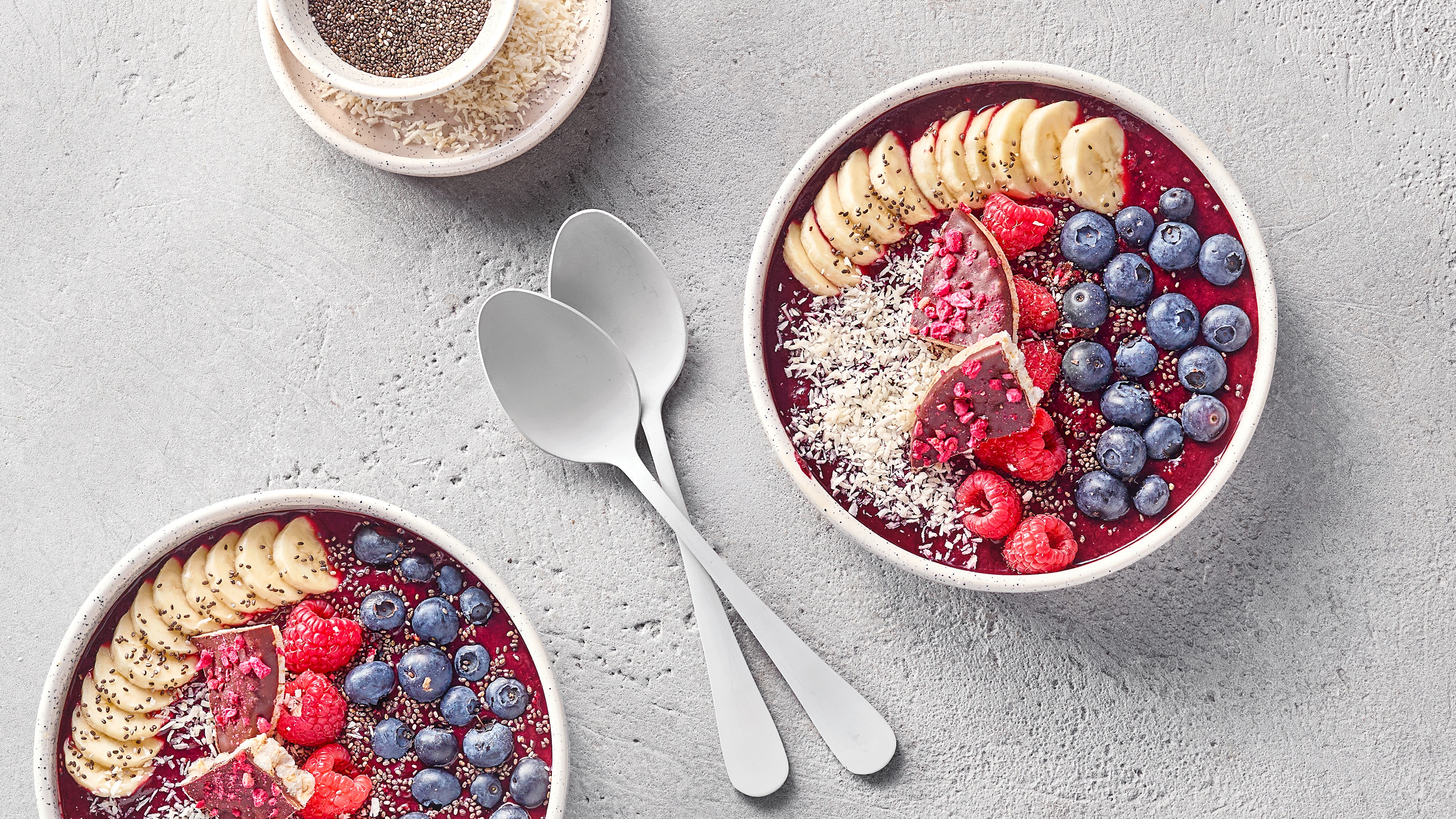 Zwei Schalen mit roter Creme, Bananenscheiben, Heidelbeeren, Himbeeren und Kokosraspeln