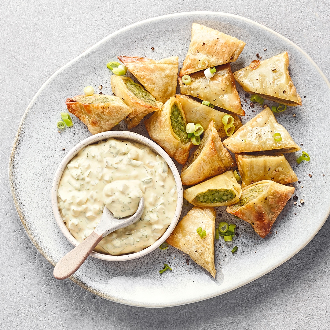 Knusprige, dreieckige Teigtaschen mit grüner Füllung auf einem Teller, serviert mit einem cremigen Dip und bestreut mit Frühlingszwiebeln.