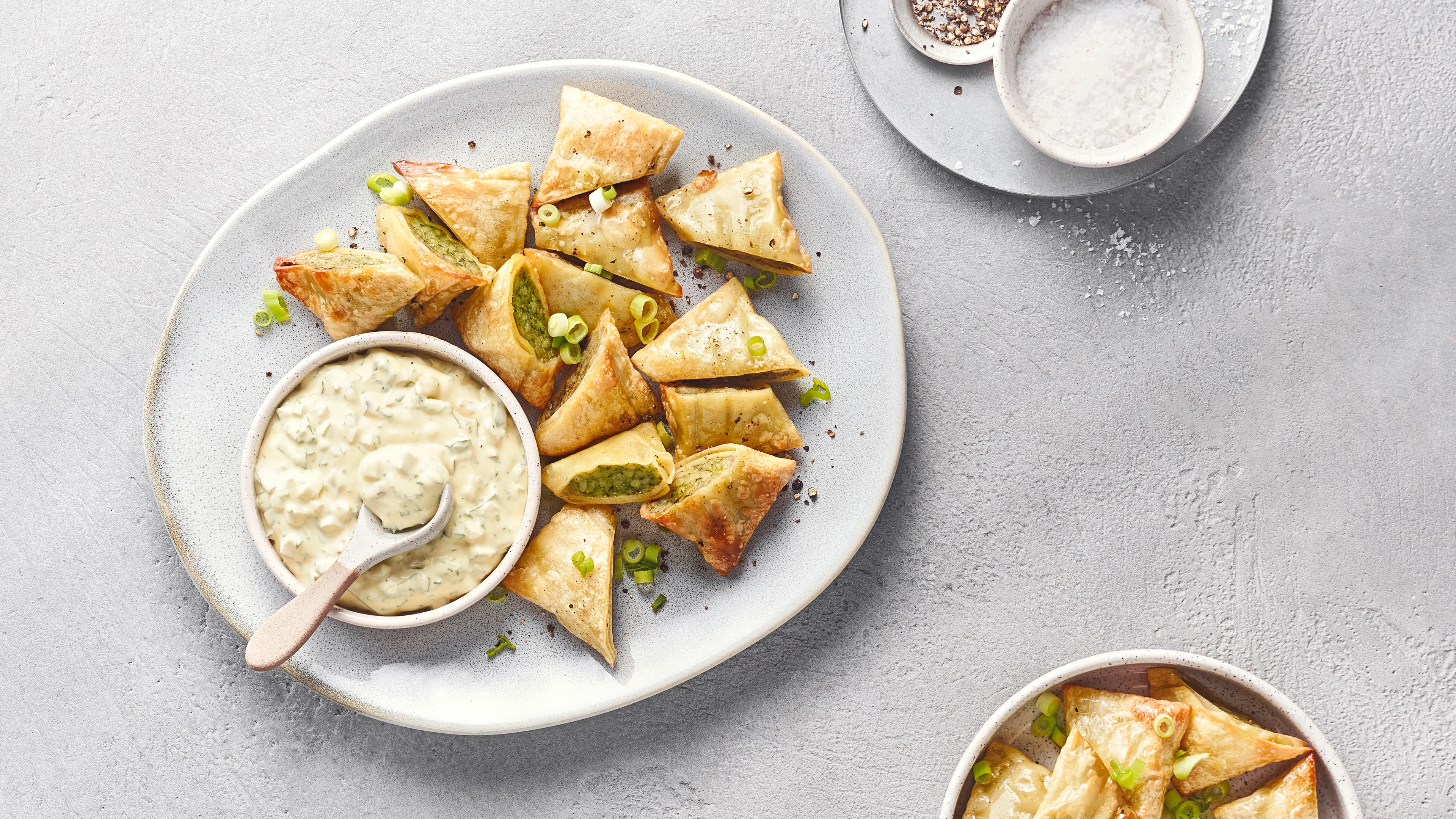Knusprige, dreieckige Teigtaschen mit grüner Füllung auf einem Teller, serviert mit einem cremigen Dip und bestreut mit Frühlingszwiebeln.