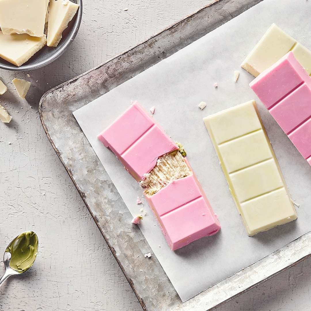 Längliche Tafeln weiße und pastellrosa Schokolade, gefüllt mit einer hellen, fädigen Creme, daneben ein Löffel mit Pistazienmus
