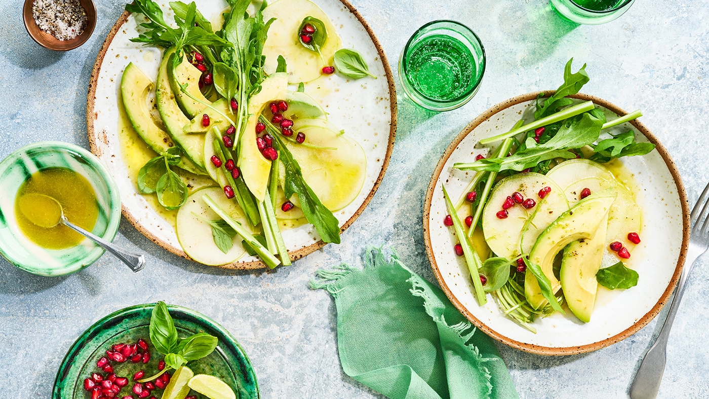 Ein großer und ein kleiner Teller mit Apfelscheiben, Avocadoscheiben, Rucolablättern, Granatapfelkernen und Dressing
