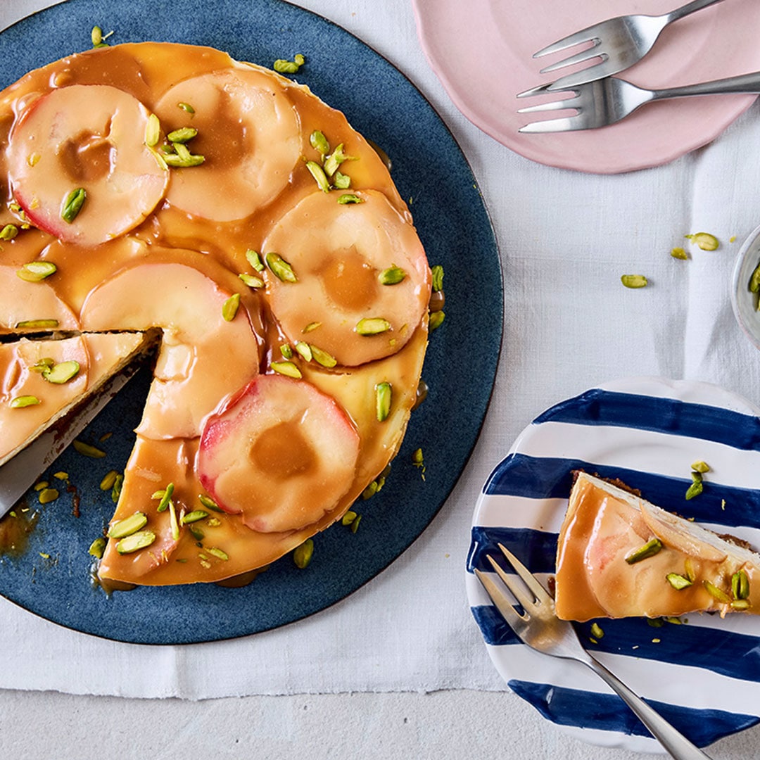 Ein runder Kuchen, getoppt mit Apfelscheiben, darauf karamellfarbene Soße und gehackte Pistazien, auf einem großen Teller