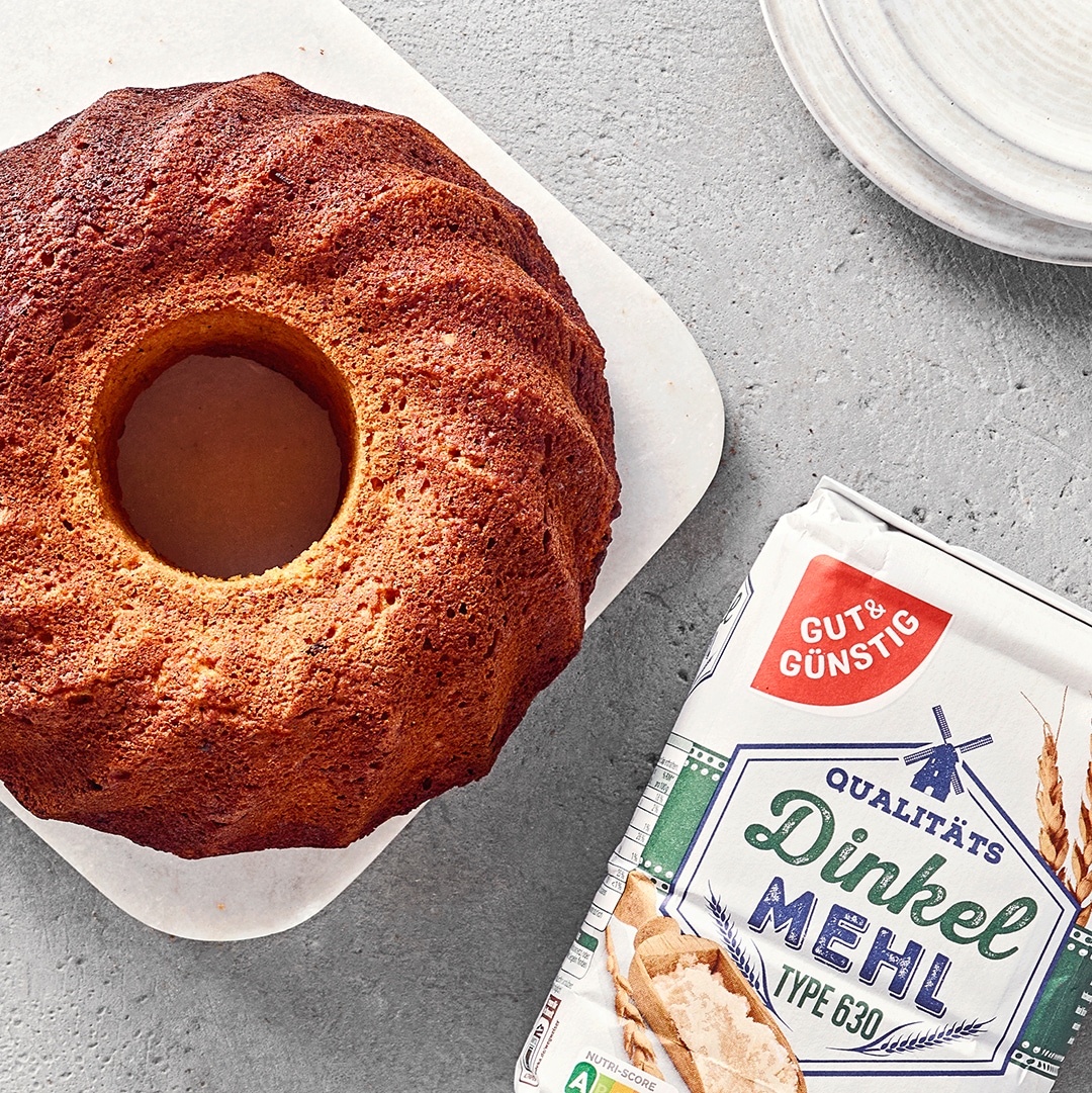 Goldbrauner Gugelhupf auf einer Servierplatte, daneben eine Packung GUT&GÜNSTIG Dinkelmehl und ein Dessertteller.