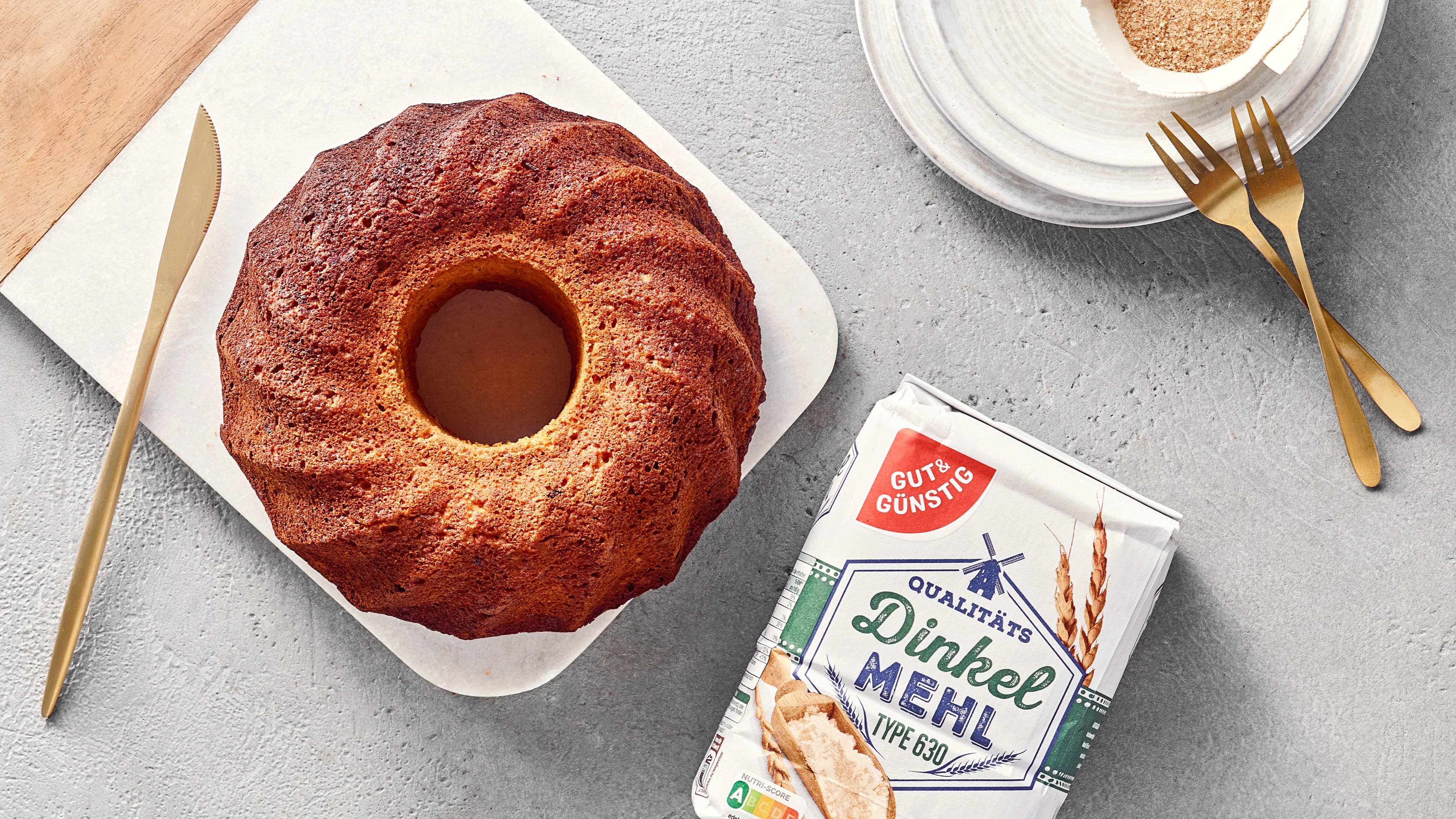 Goldbrauner Gugelhupf auf einer Servierplatte, daneben eine Packung GUT&GÜNSTIG Dinkelmehl sowie goldfarbenes Besteck und ein Dessertteller.