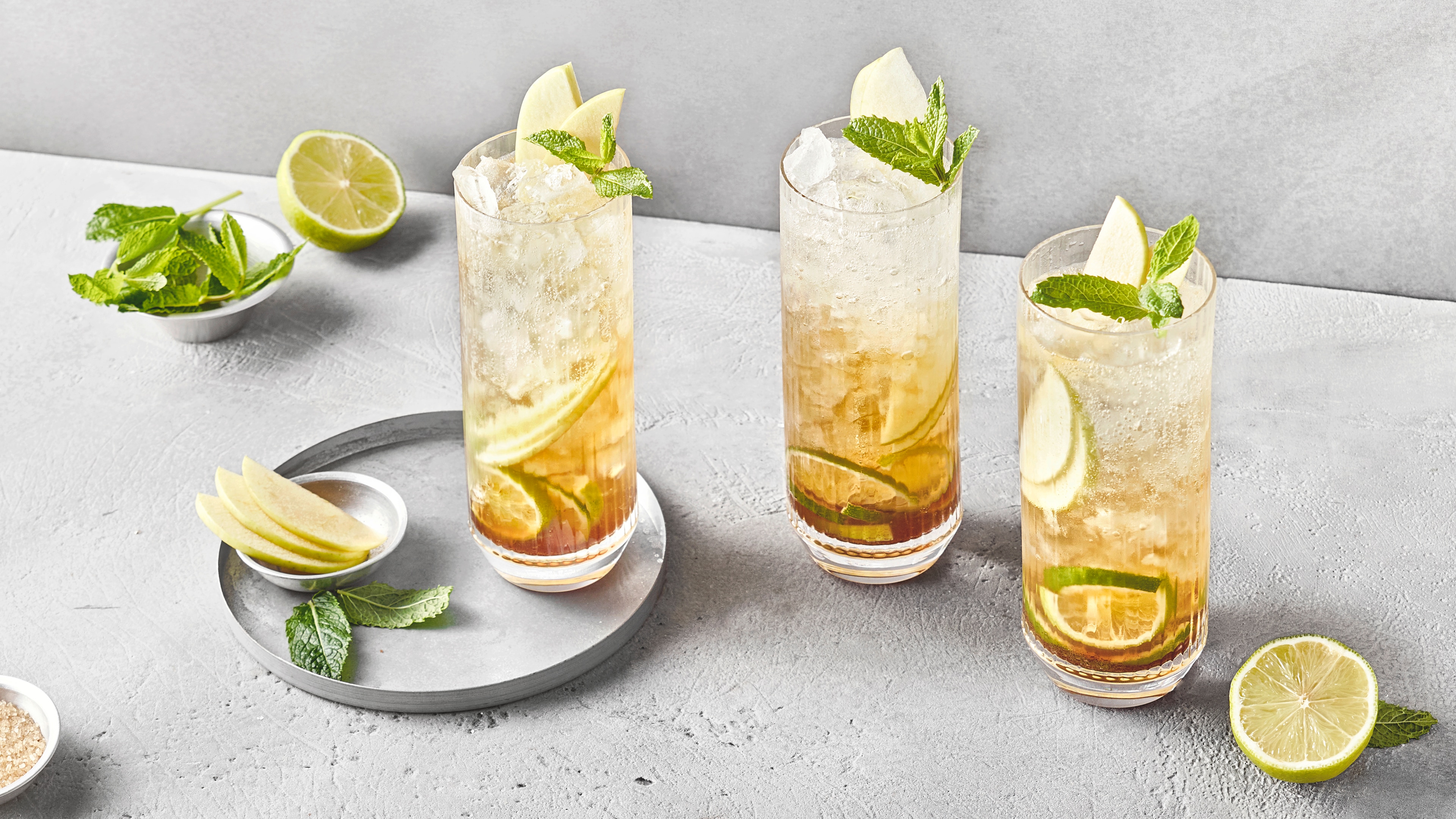 Drei Cocktailgläser mit Äpfeln, Limetten- und Zitronenscheiben, Eiswürfeln und Minzblättern.