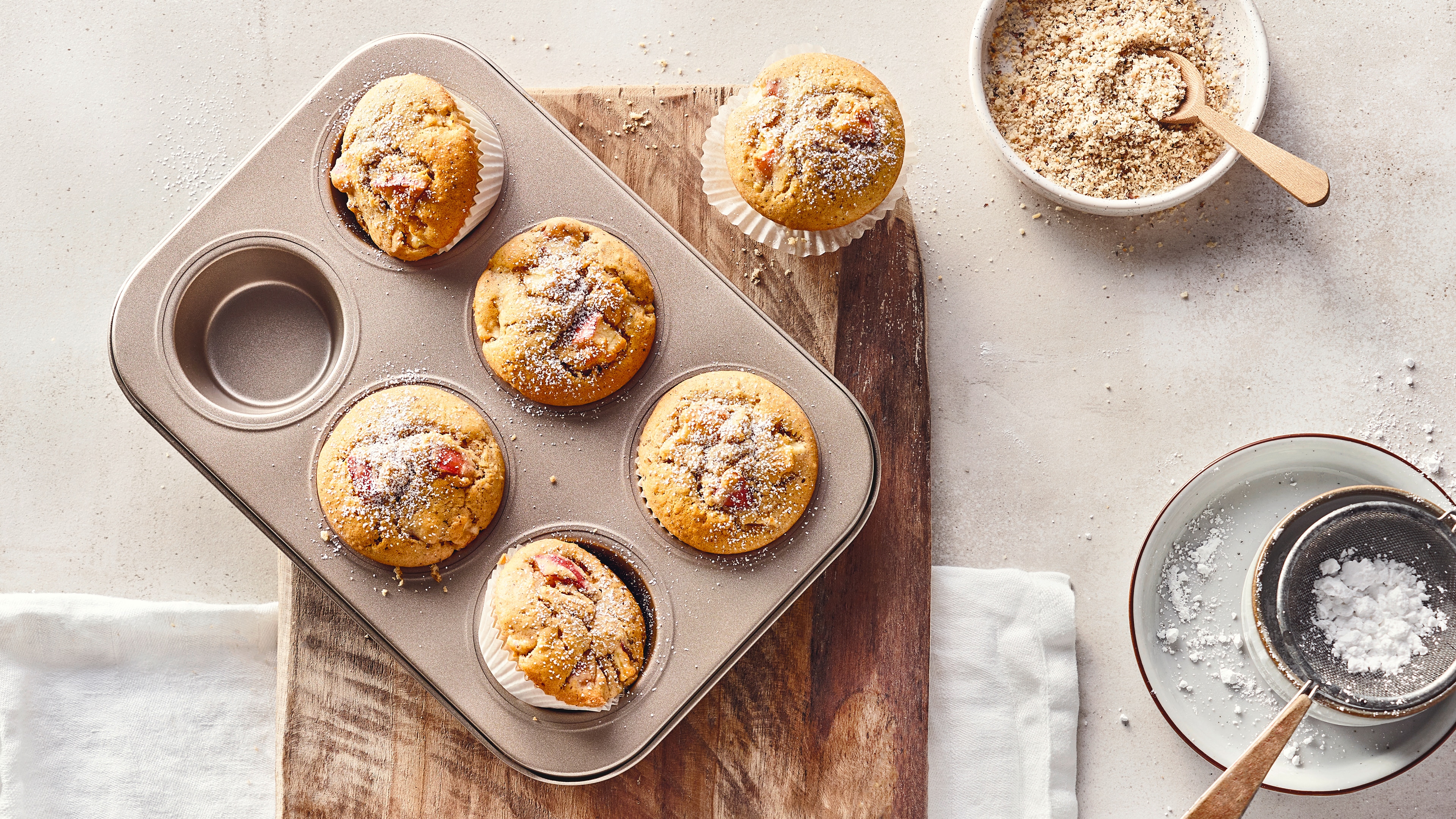 Rezept für Apfel-Nuss-Muffins Eine Muffin-Backform mit hellen, fluffigen Muffins mit Apfelstückchen, bestreut mit Puderzucker