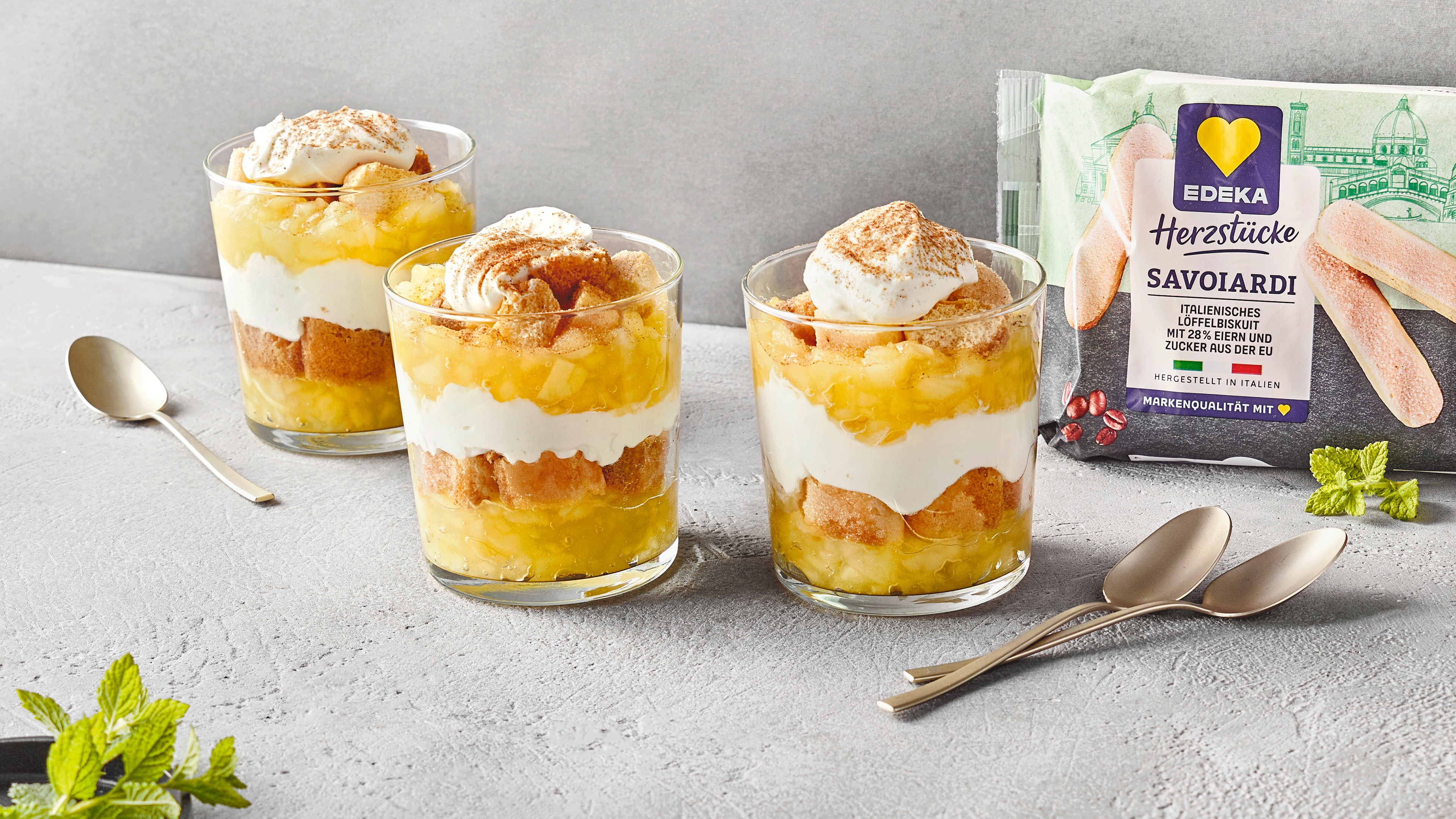 Drei Schichtdessertgläser mit abwechselnder Apfelschicht und weißer Creme. Rechts daneben EDEKA Herzstücke Savoiardi (Löffelbiscuits).