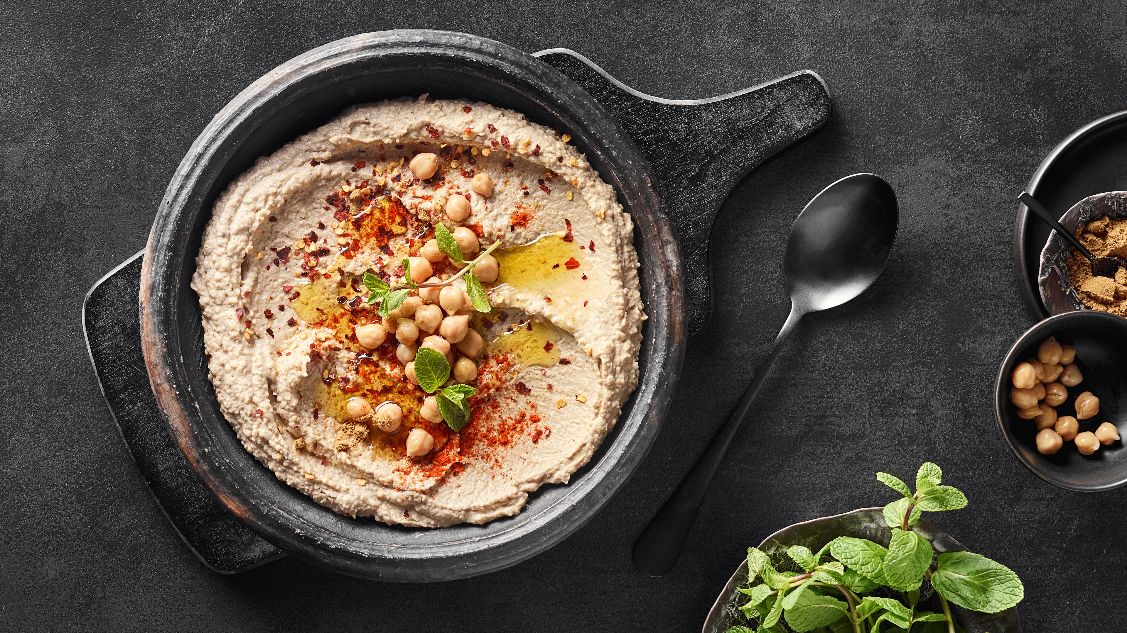 Eine Schüssel mit beigem Püree, darauf helle Hülsenfrüchte, grüne Blätter, rote Flocken und gelbem Öl.