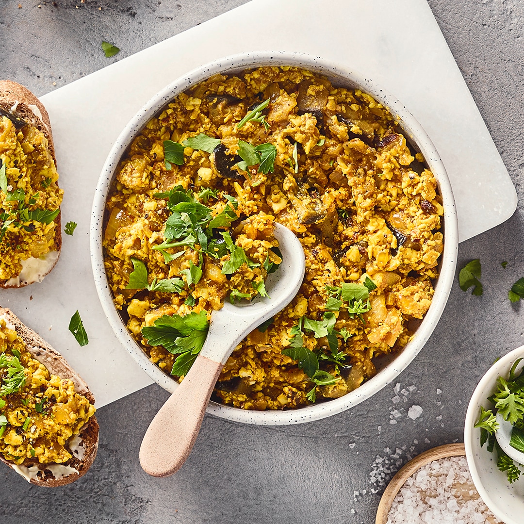 Eine Schüssel mit gelber krümeliger Masse, grünen Kräutern und dunklen Würfeln, daneben zwei Brotscheiben.